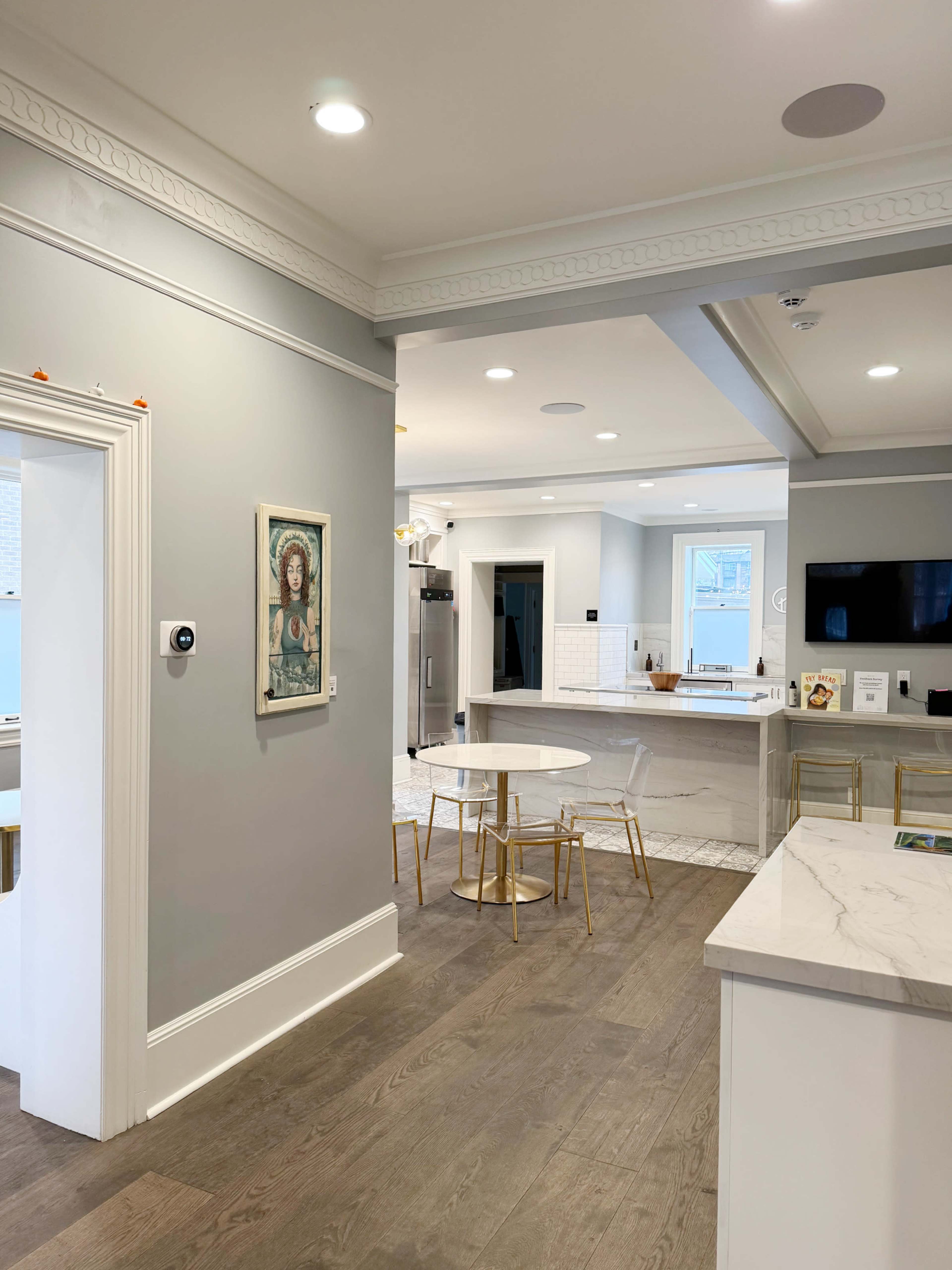 A modern kitchen features a gray wall with a framed artwork, a dining area with a small round table and gold chairs, and a seamless flow into a well-lit living space.