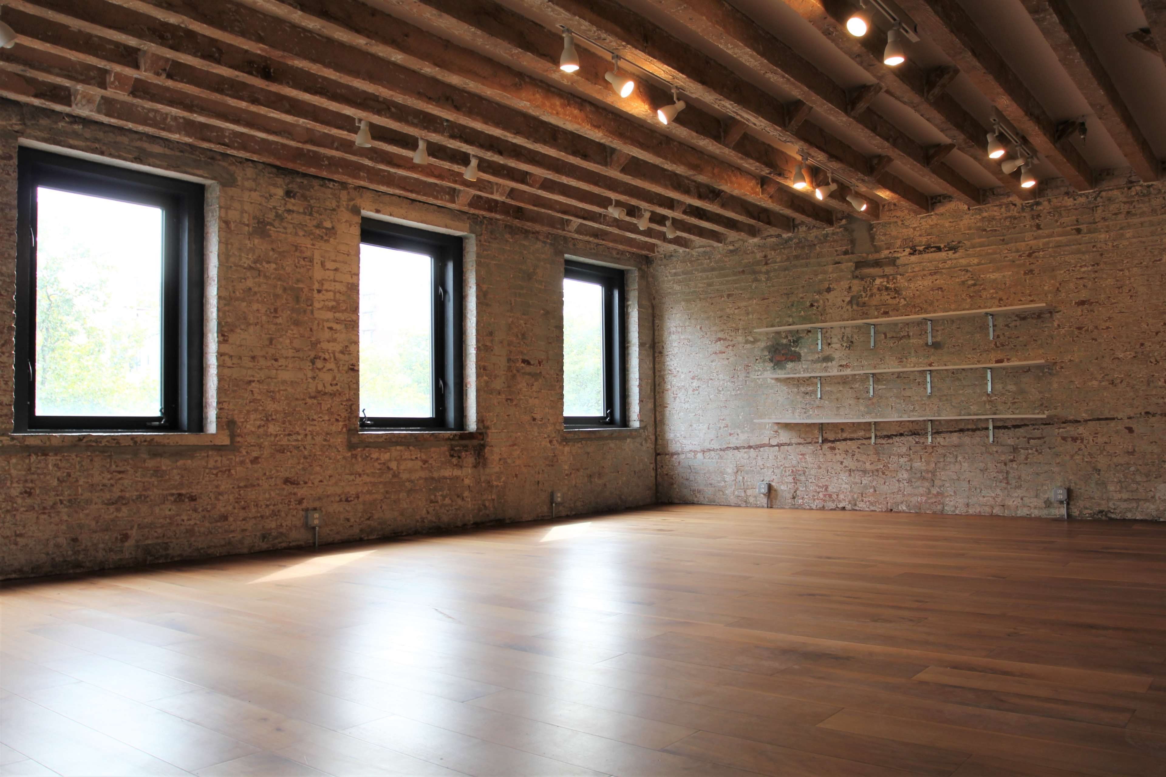 A spacious interior room with exposed brick walls, wooden floors, and large windows illuminating the space.