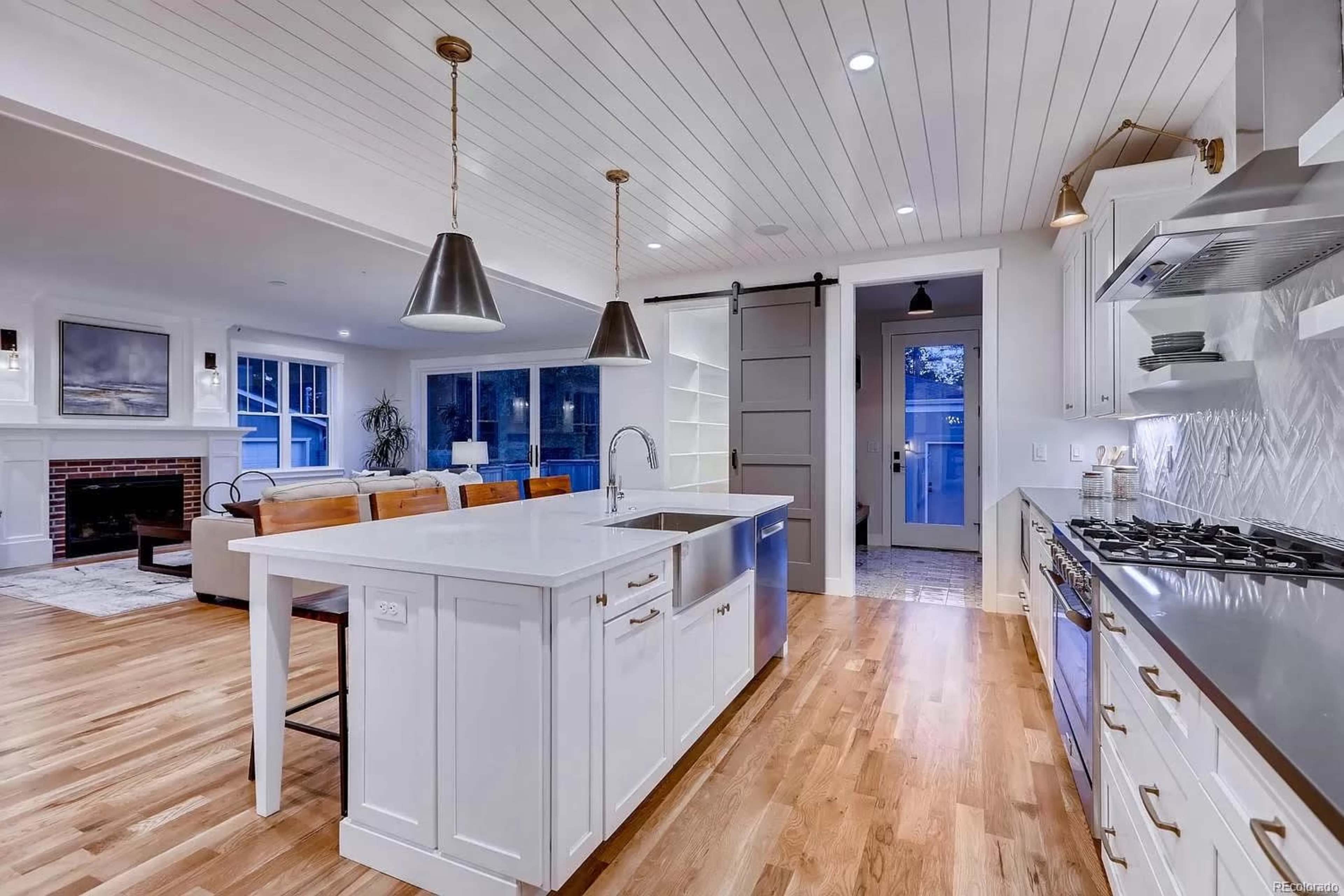 The image shows a modern kitchen with a large island, stainless steel appliances, pendant lighting, and a living area in the background featuring a sofa and a fireplace.