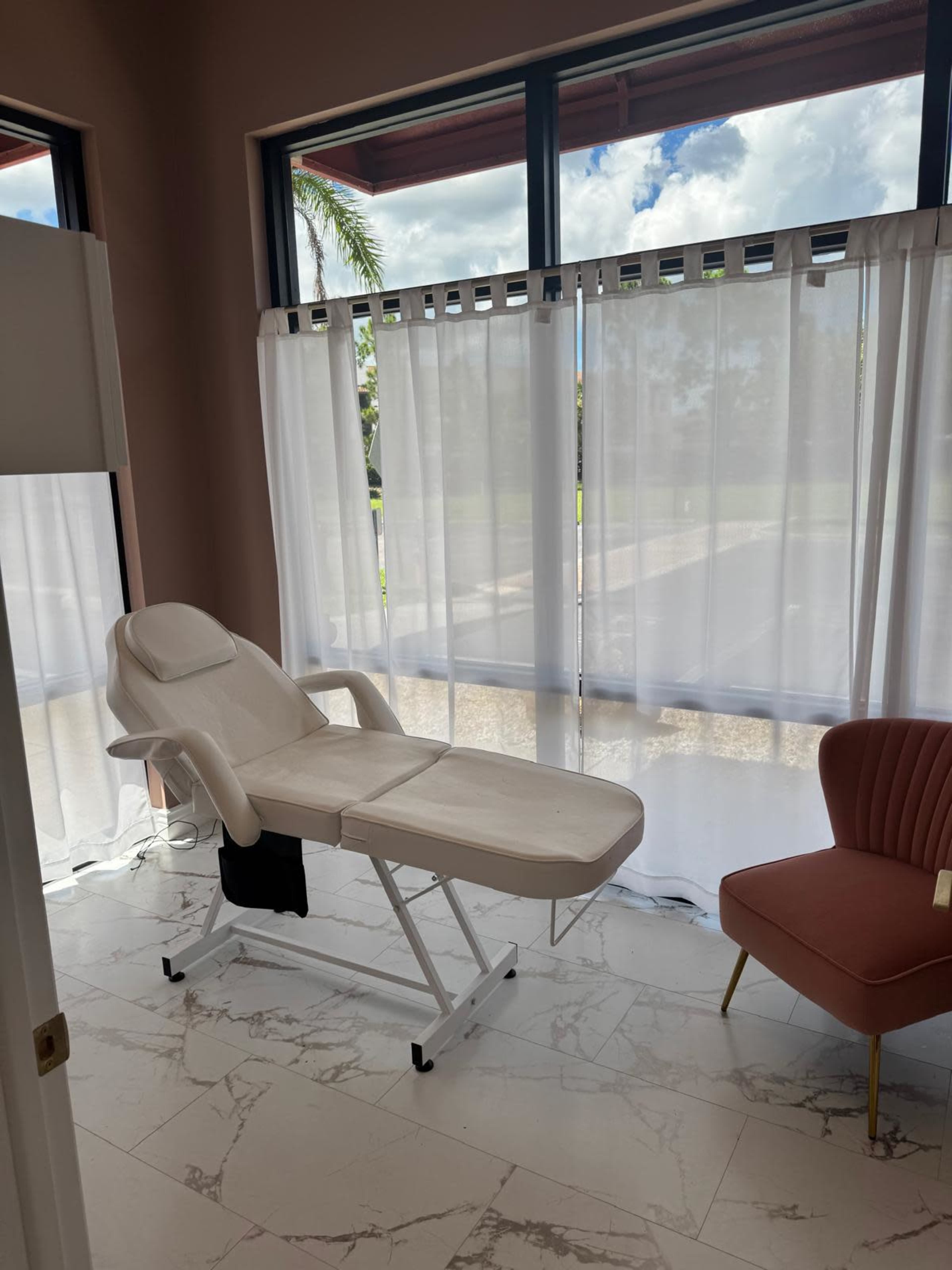 The image shows a bright, airy room with a treatment table, a pink accent chair, and sheer curtains letting in natural light.