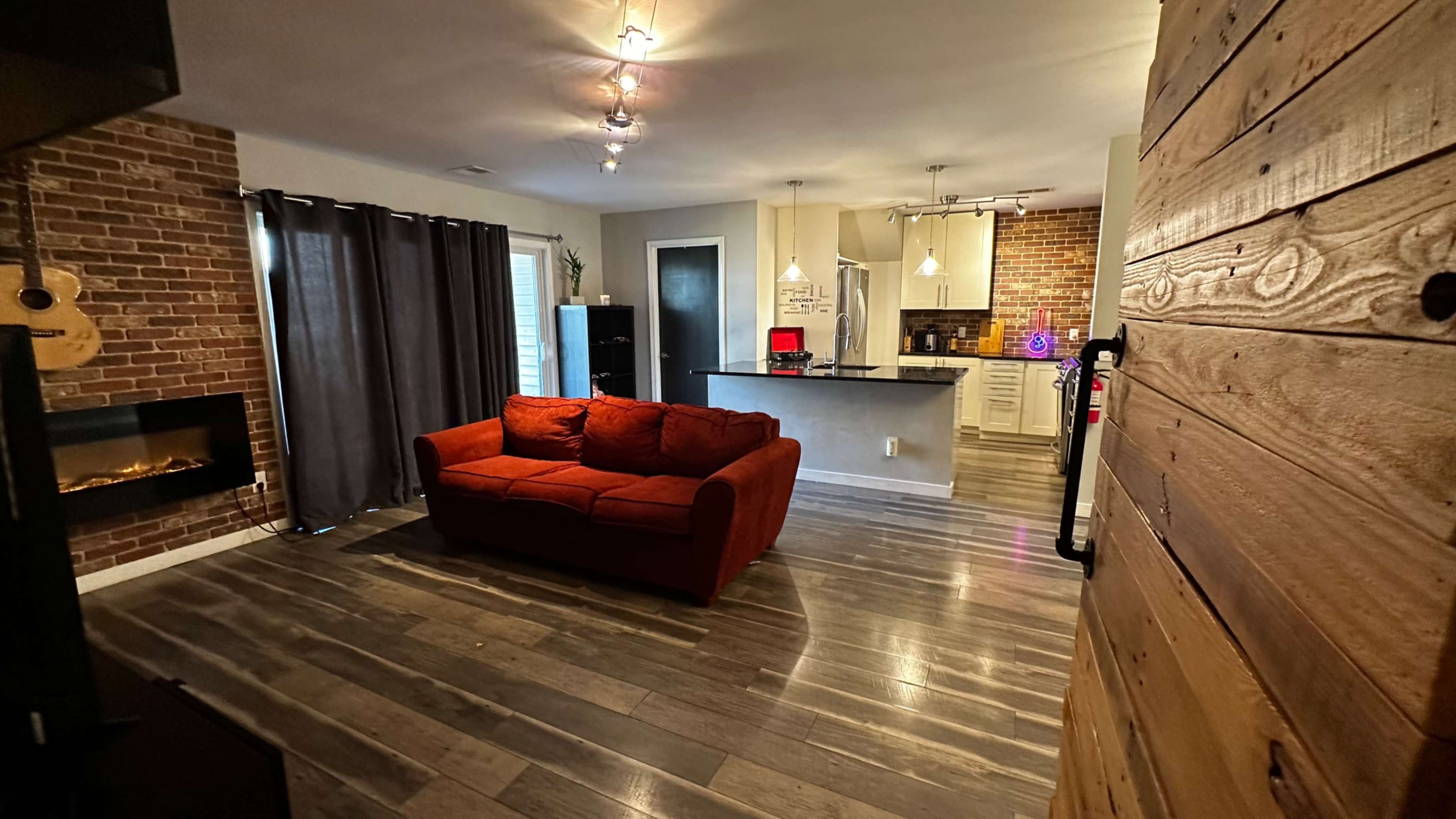 A living room with a red sofa, a brick wall, dark curtains, and an open kitchen area in the background.