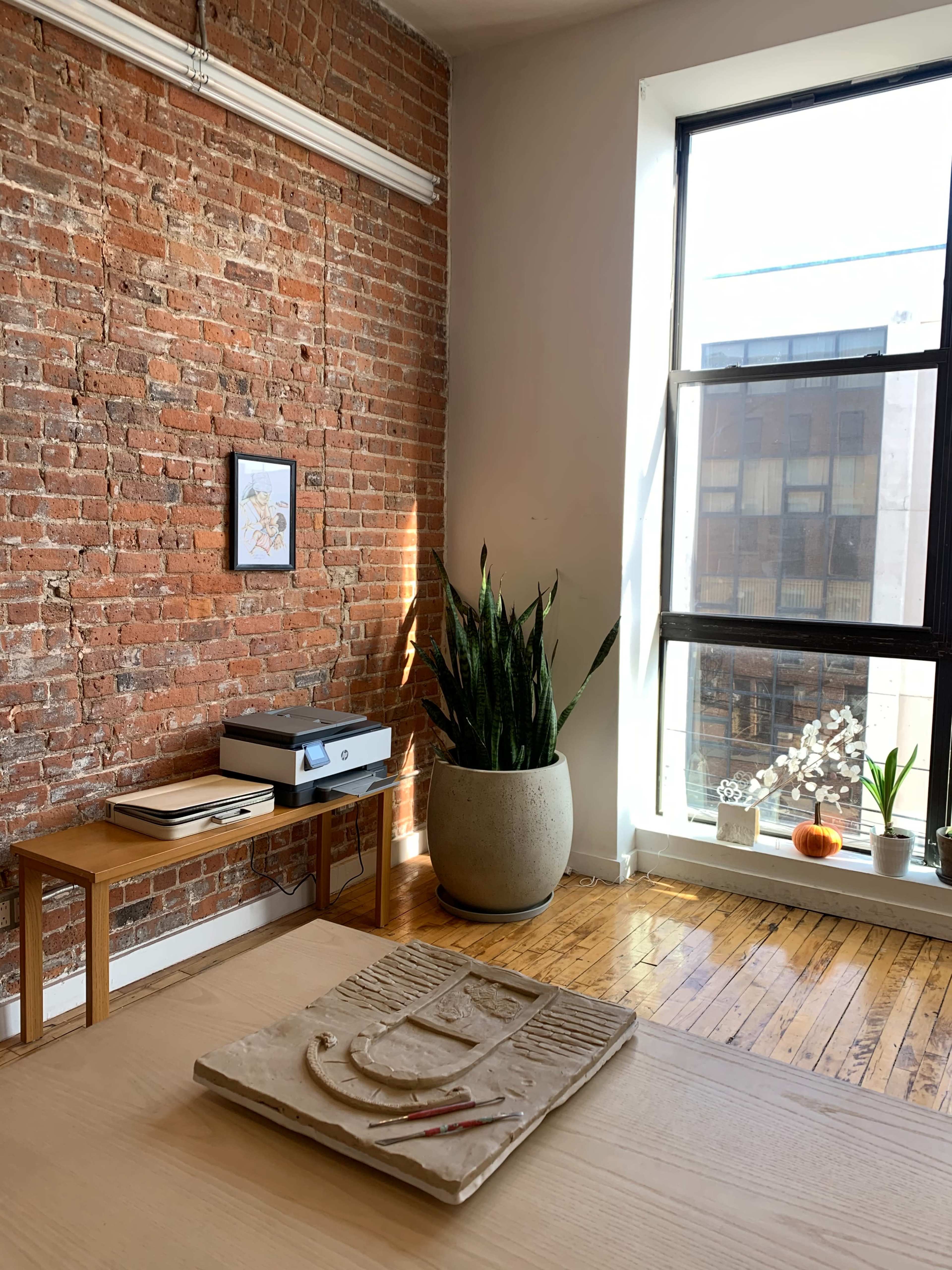 The image shows a minimalist workspace with a wooden table at the center, a brick wall, plants in the corner, and a window letting in natural light.