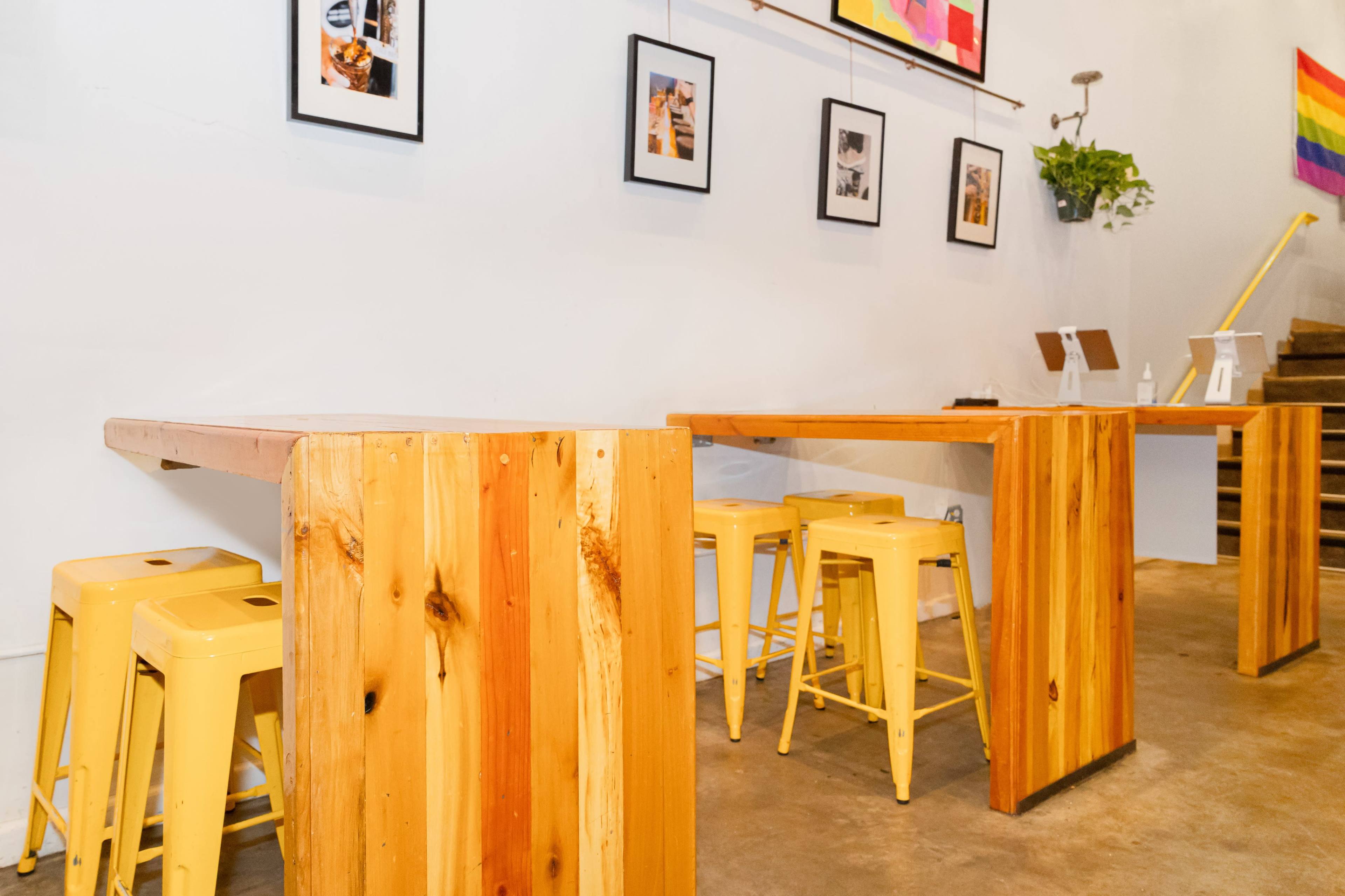 A cafe interior with wooden tables and yellow metal stools against a white wall adorned with framed pictures and a rainbow flag.