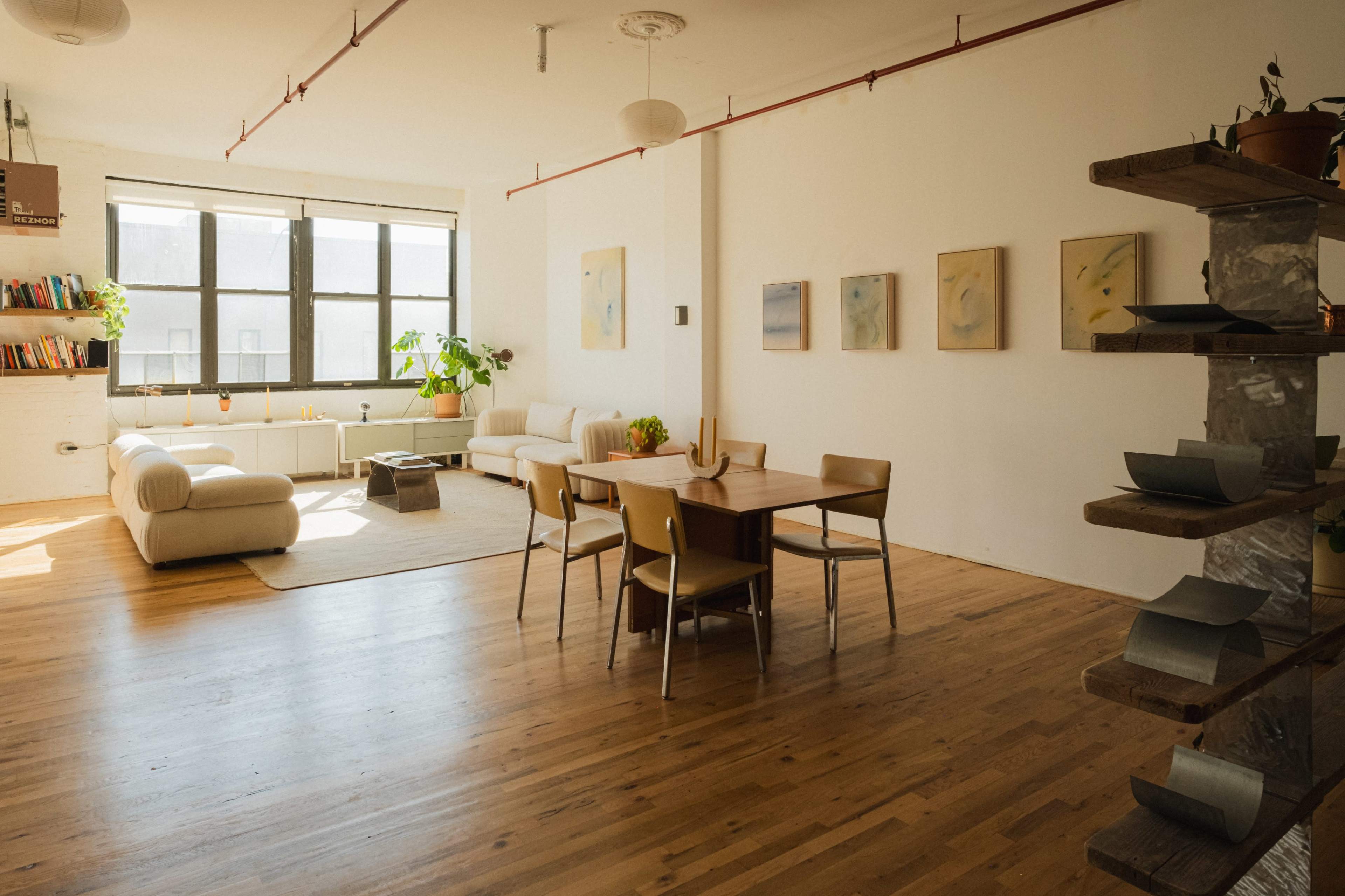 The image shows a bright, spacious living area with wooden floors, a dining table, a couch, and plants near large windows.