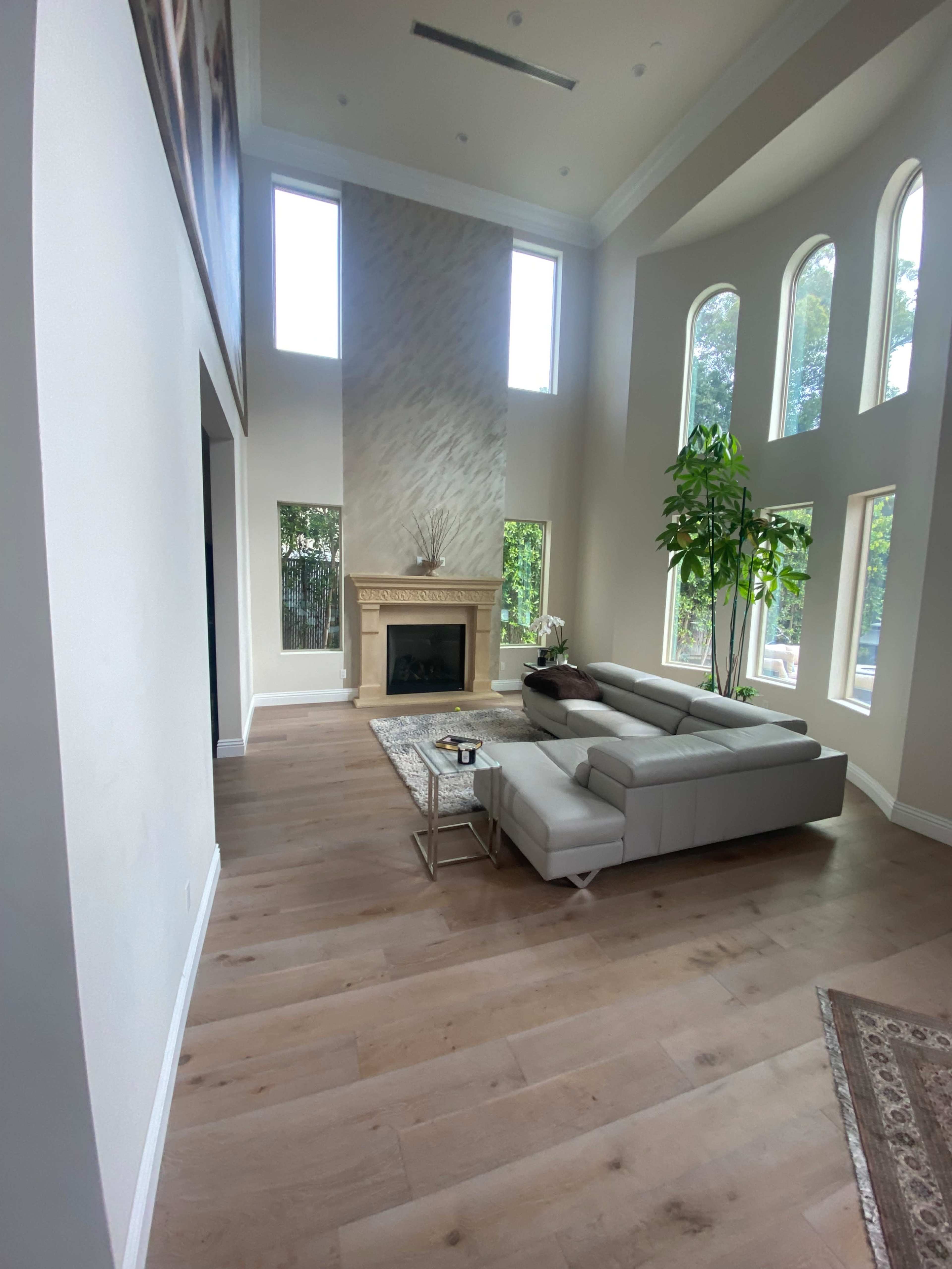 A spacious living room featuring a light-colored sectional sofa, a fireplace, large windows, and a potted plant.