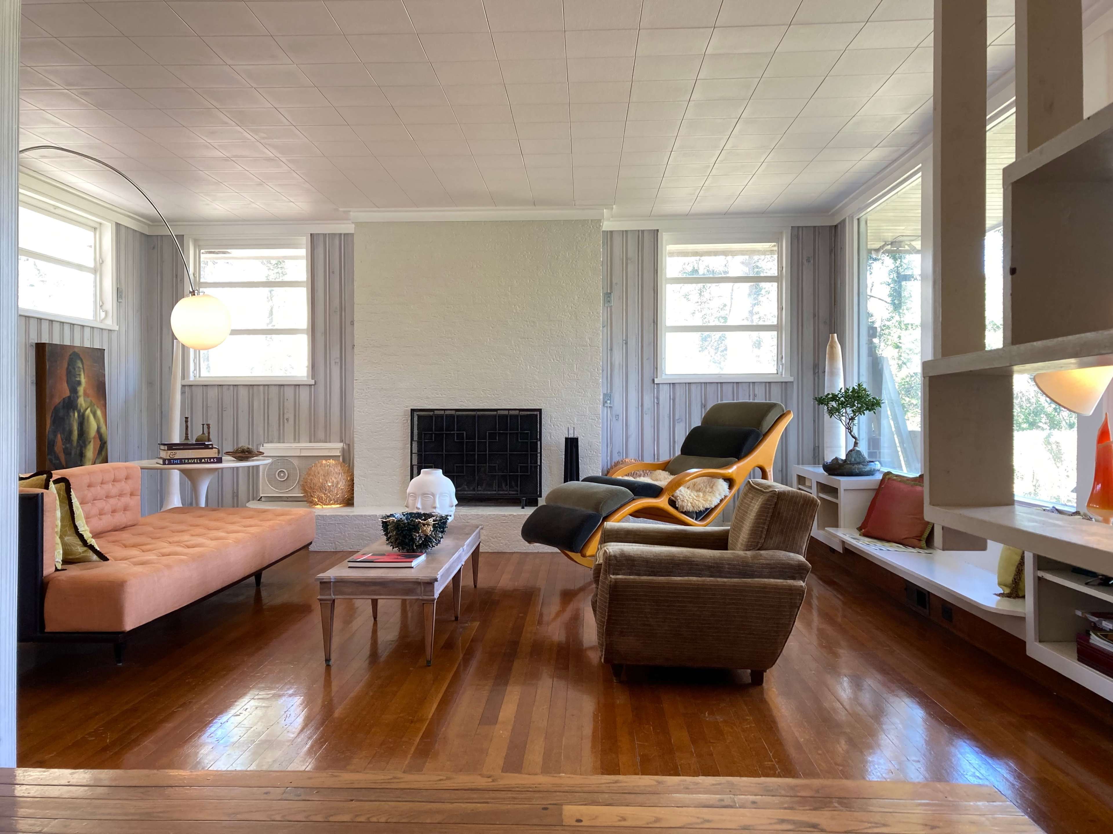 A spacious living room with a beige couch, an orange lounge chair, a wooden coffee table, and large windows allowing natural light to enter.