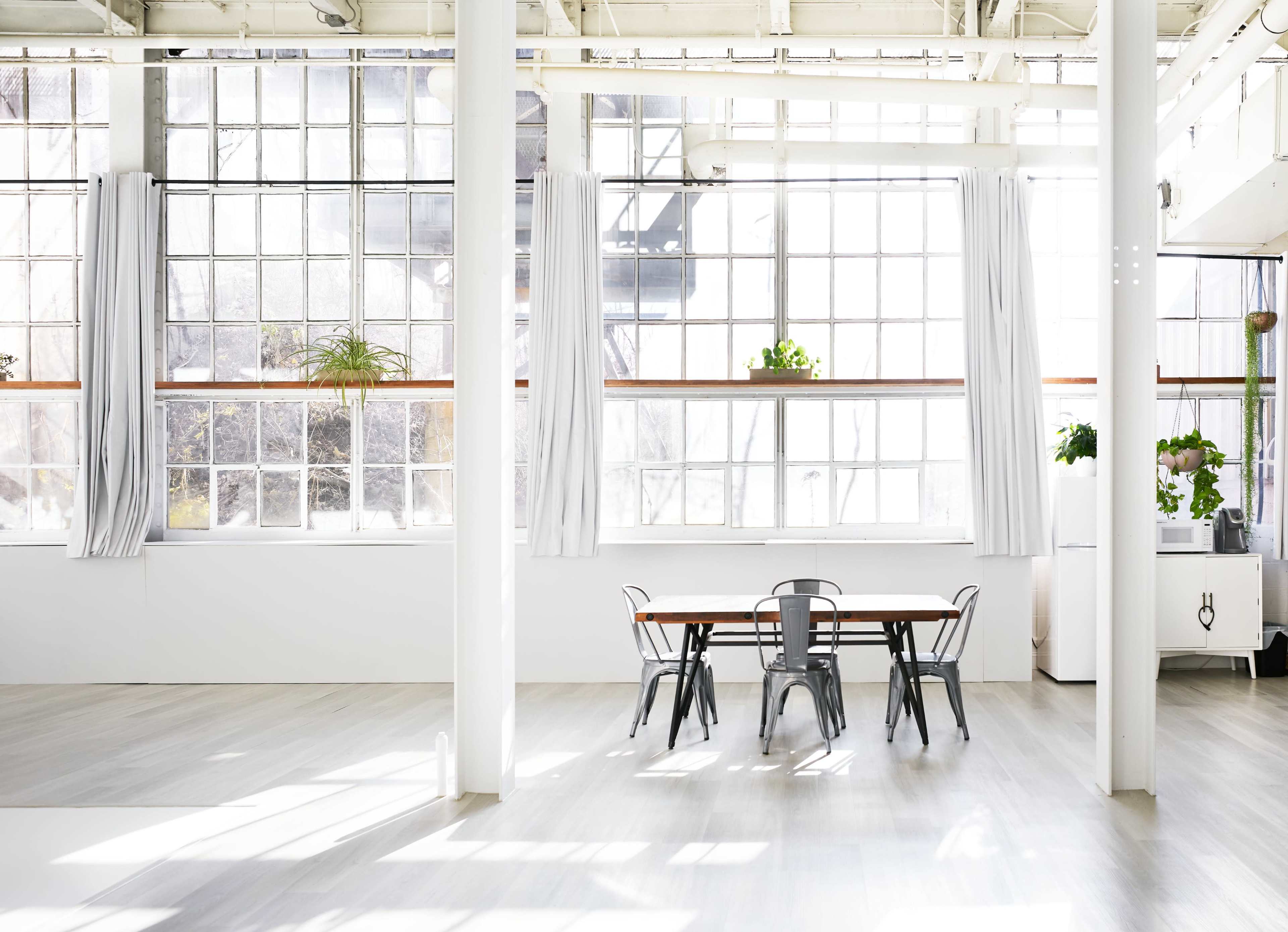 A spacious room features a long wooden table surrounded by metal chairs, with large windows allowing natural light to fill the space.