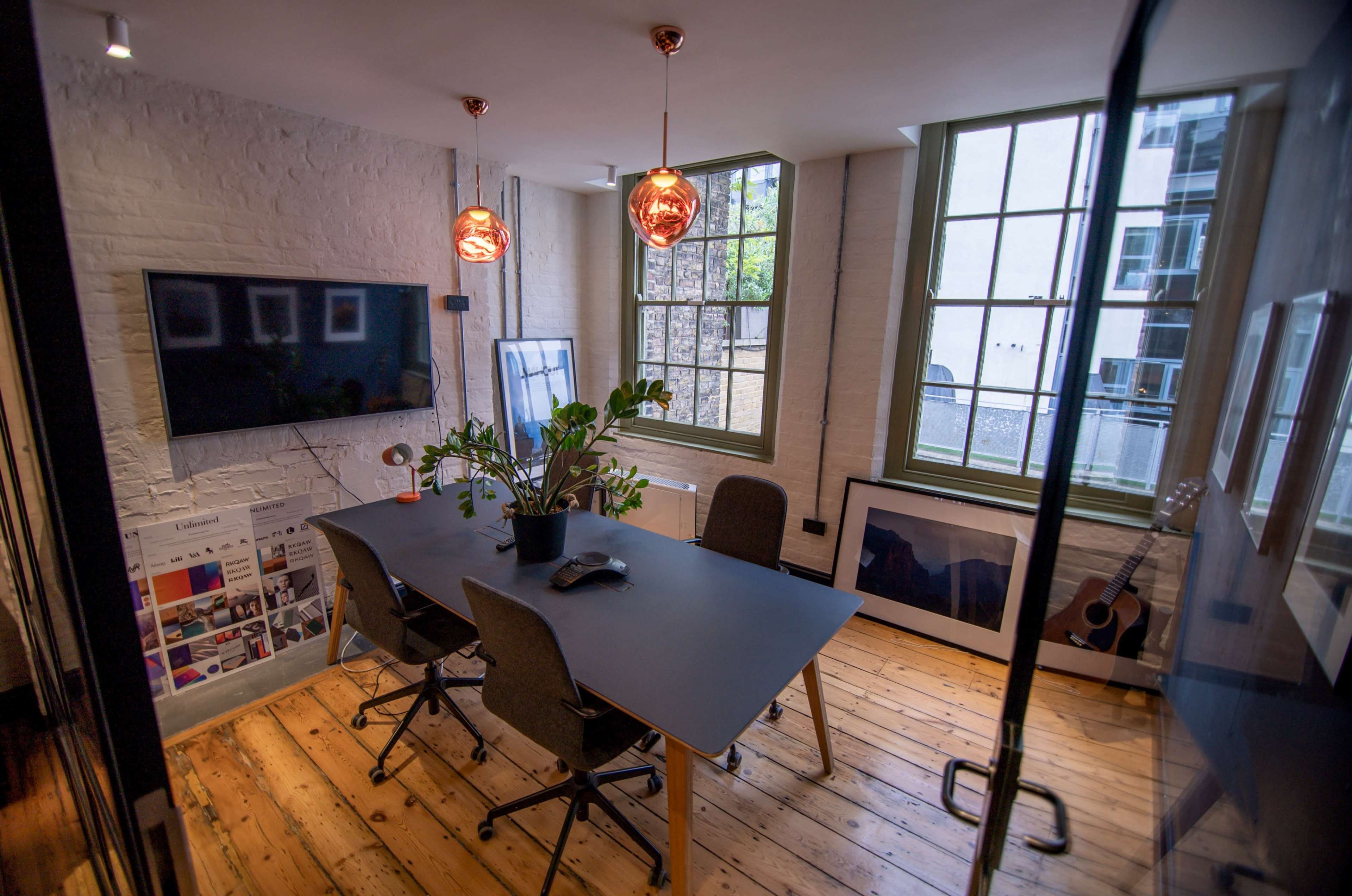 A modern office meeting room features a large table surrounded by four chairs, with a television on the wall and two hanging pendant lights above, illuminated by natural light from the windows.