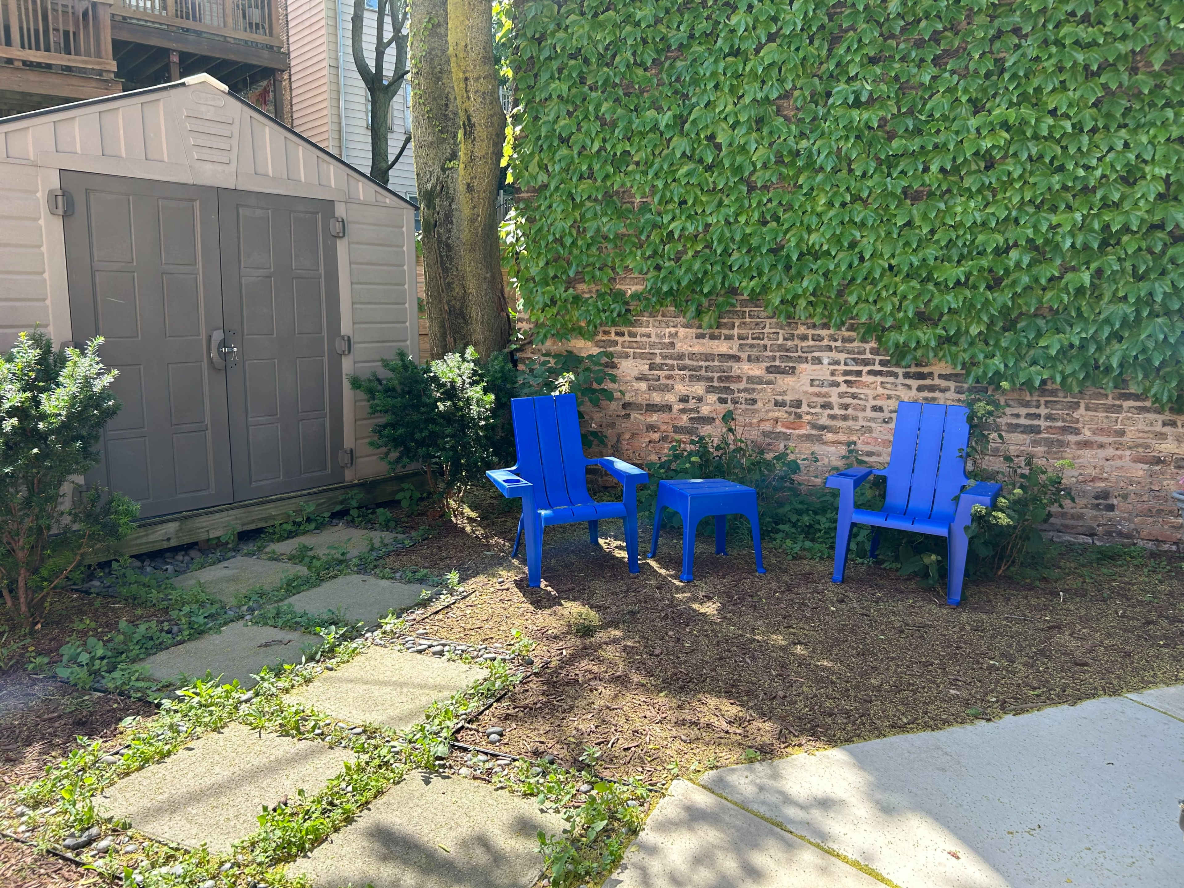 The image shows a small outdoor space with two blue chairs and a side table situated next to a storage shed, surrounded by greenery and a brick wall.