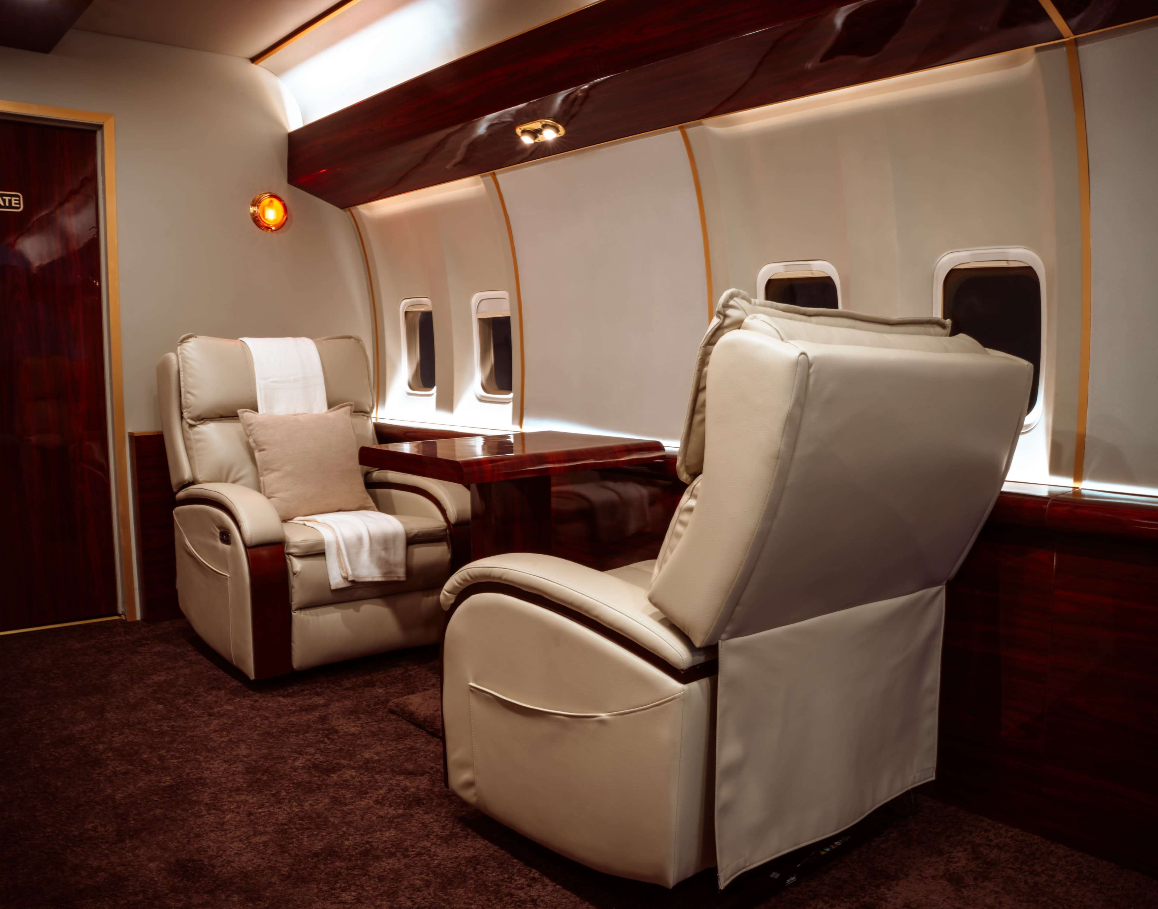 The interior of a private jet features two cream-colored recliners and a small table, with large windows allowing natural light to enter.