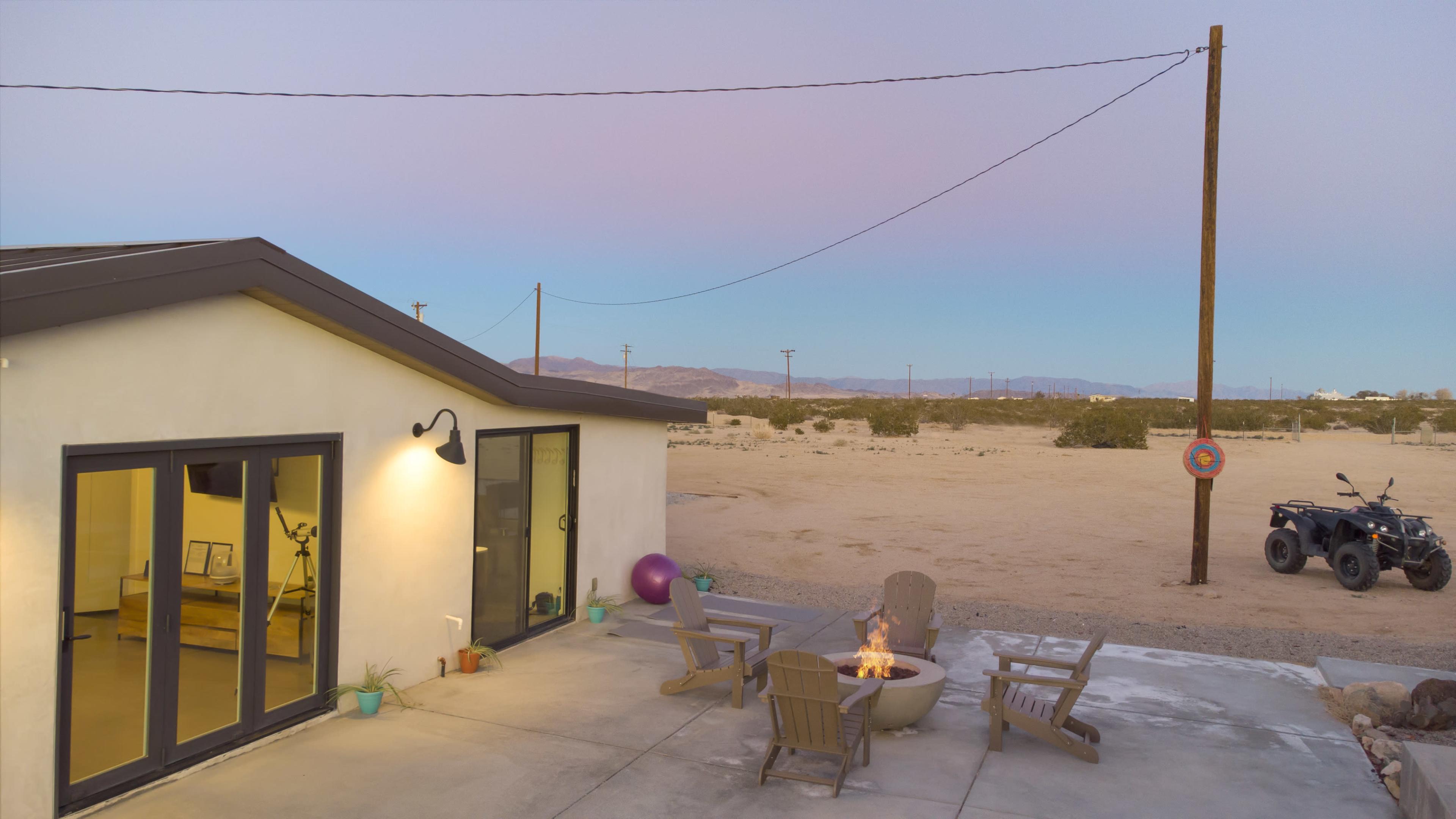 A desert home features a patio with a fire pit, outdoor seating, and distant mountains under a twilight sky.