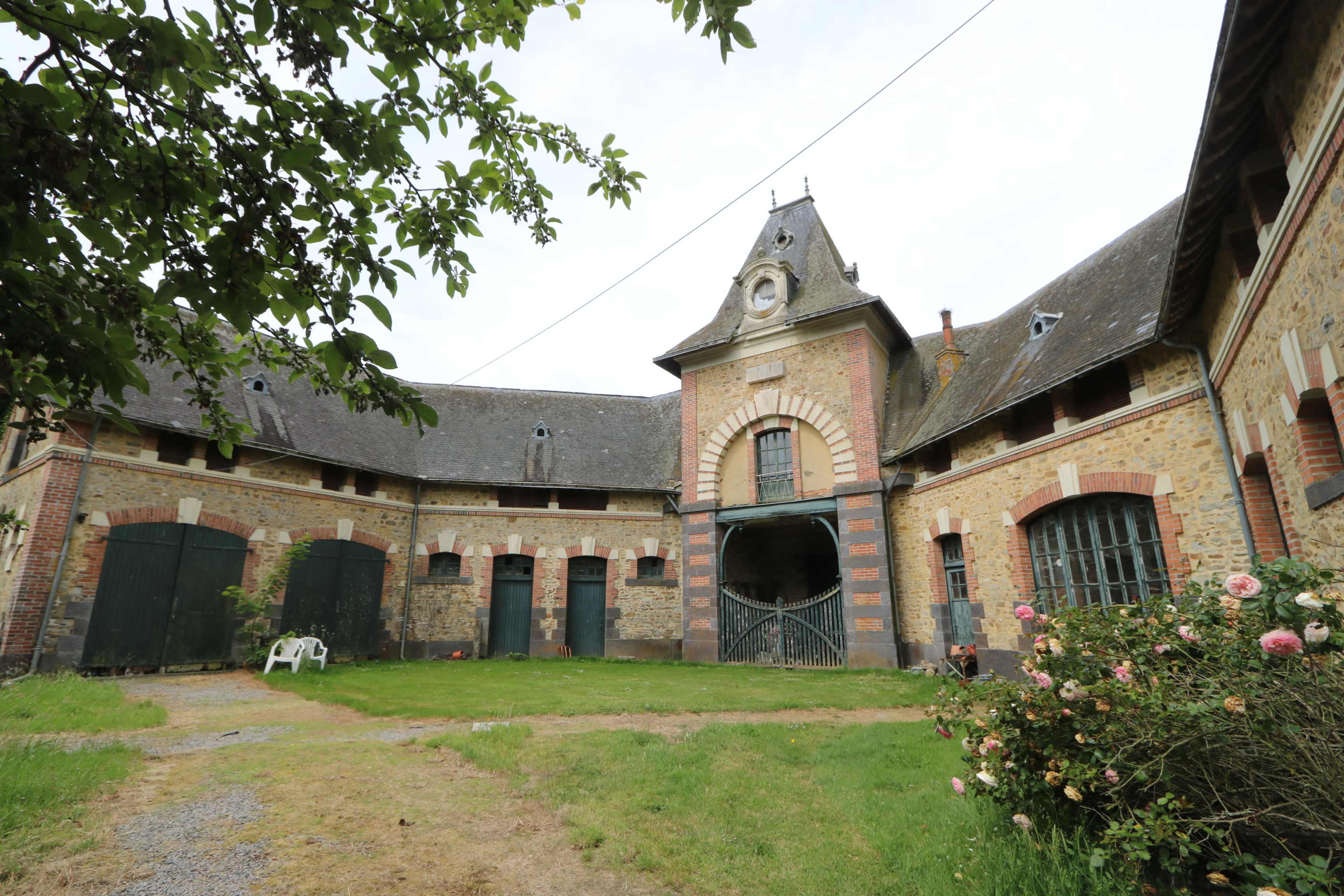 The image features a spacious courtyard surrounded by a historic brick building with a clock tower, flanked by greenery and blooming roses.