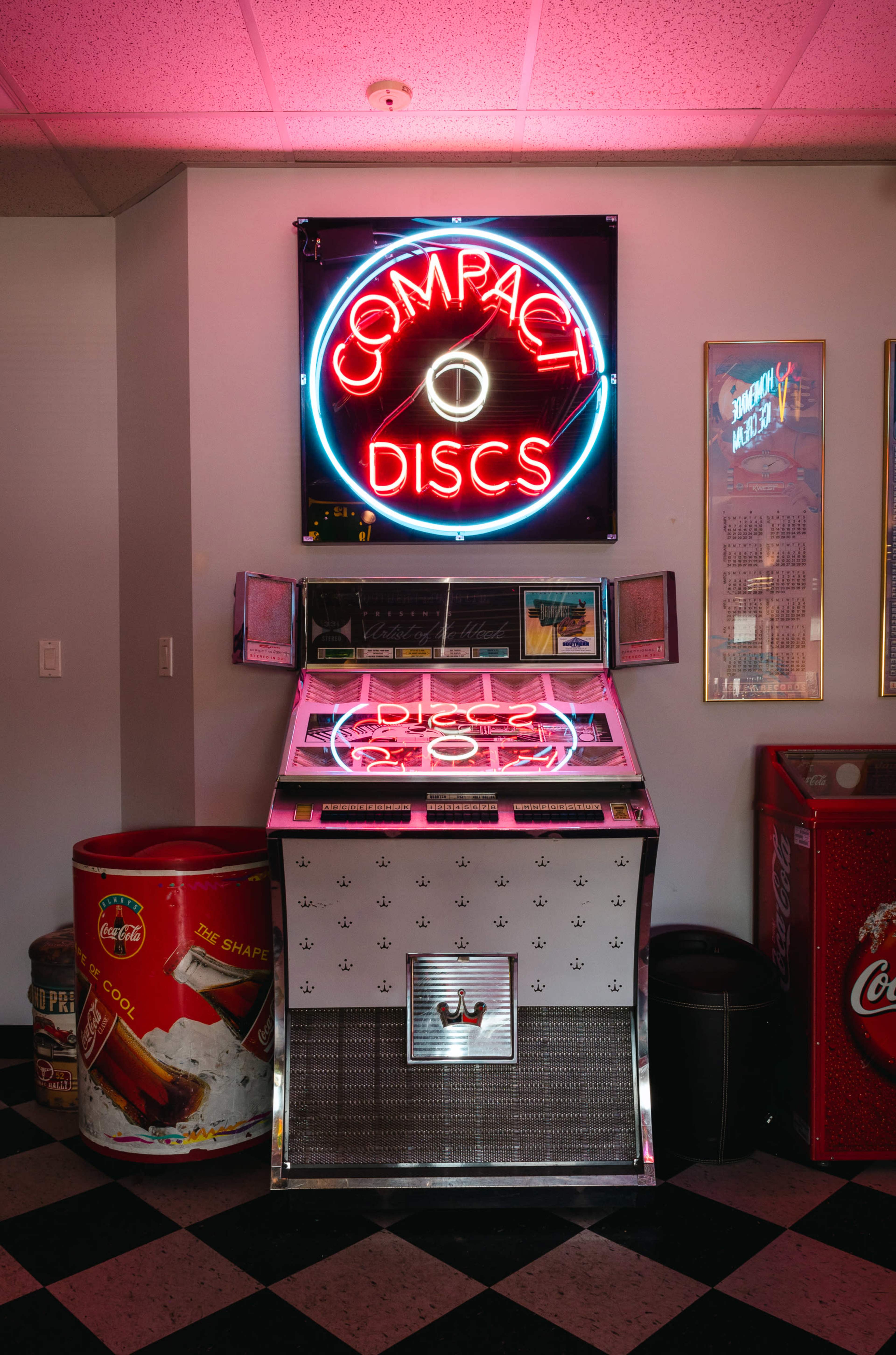 A vintage jukebox featuring a neon sign that reads "COMPACT DISCS" stands against a wall in a room with black and white checkered flooring.