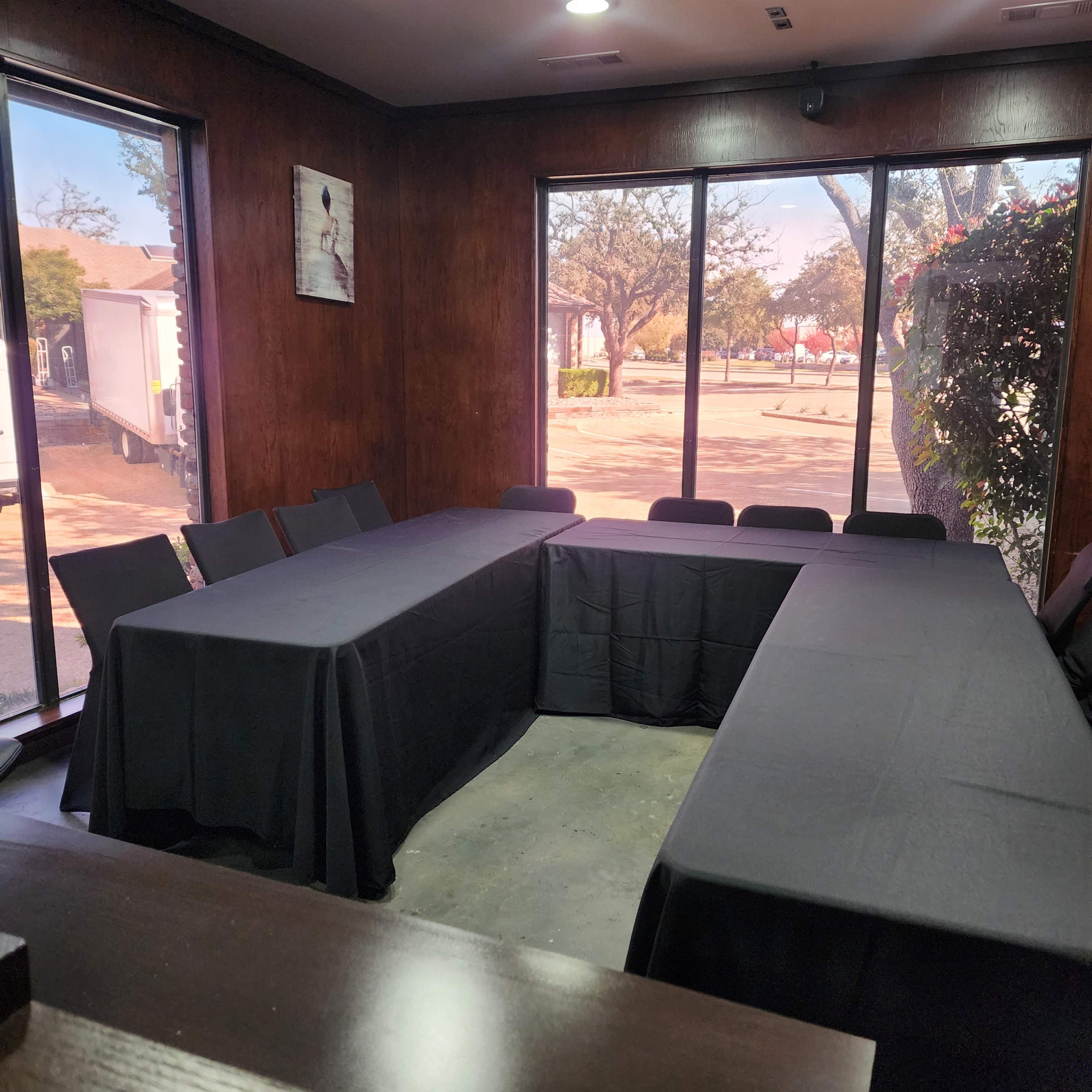 A meeting room features several black tablecloth-covered tables arranged in a U-shape, with large windows offering views of the outside.