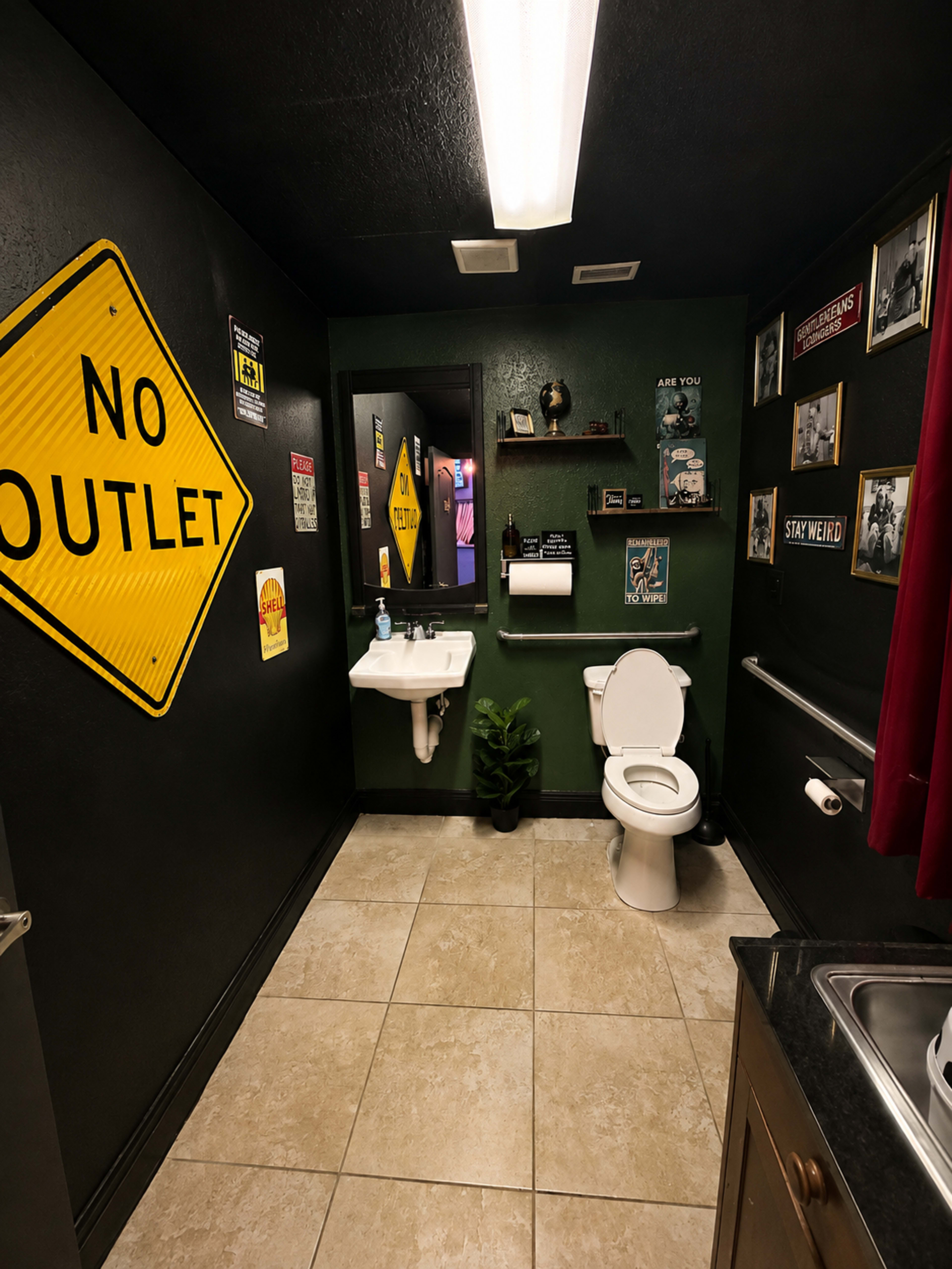 The image shows a small bathroom with a dark green wall, a white sink, a toilet, a variety of wall signs, and a red curtain.