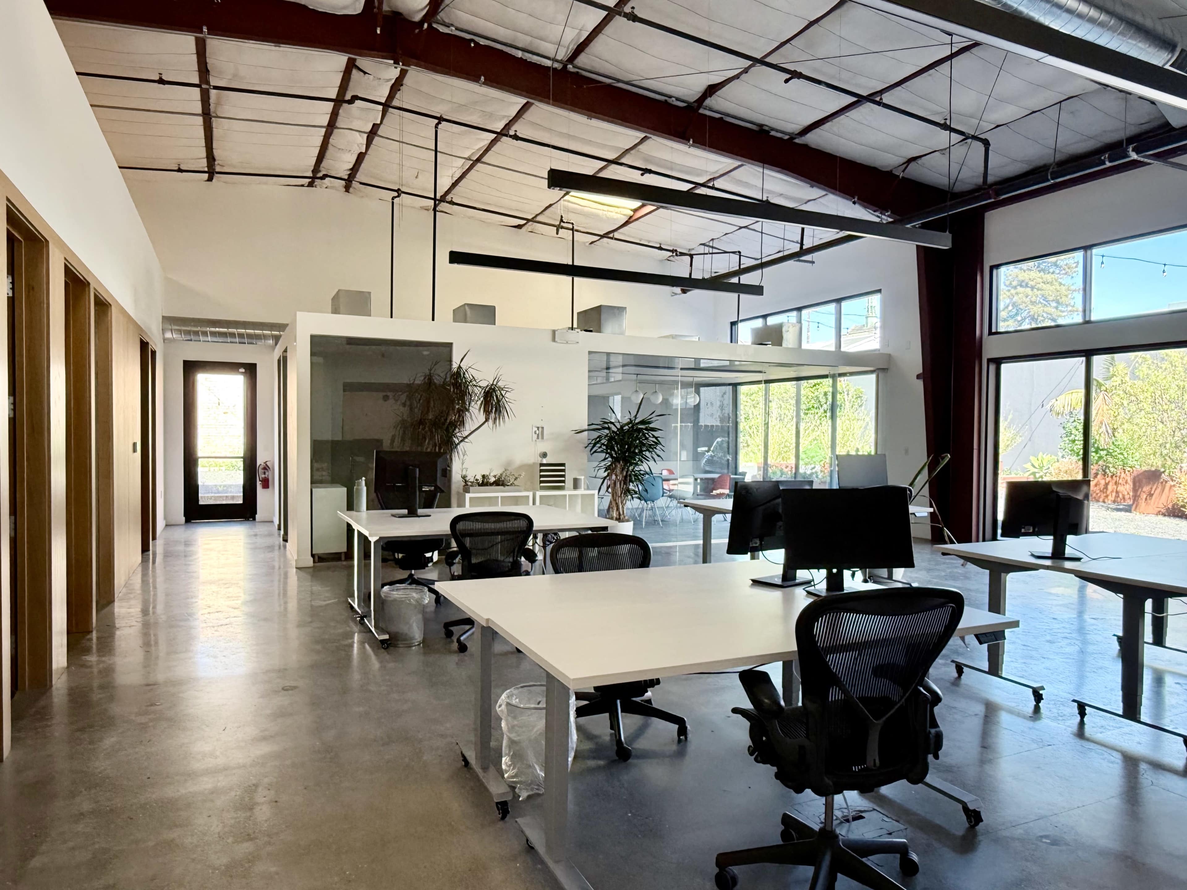 The image shows a modern, open office space with several desks, large windows, and a glass meeting room.