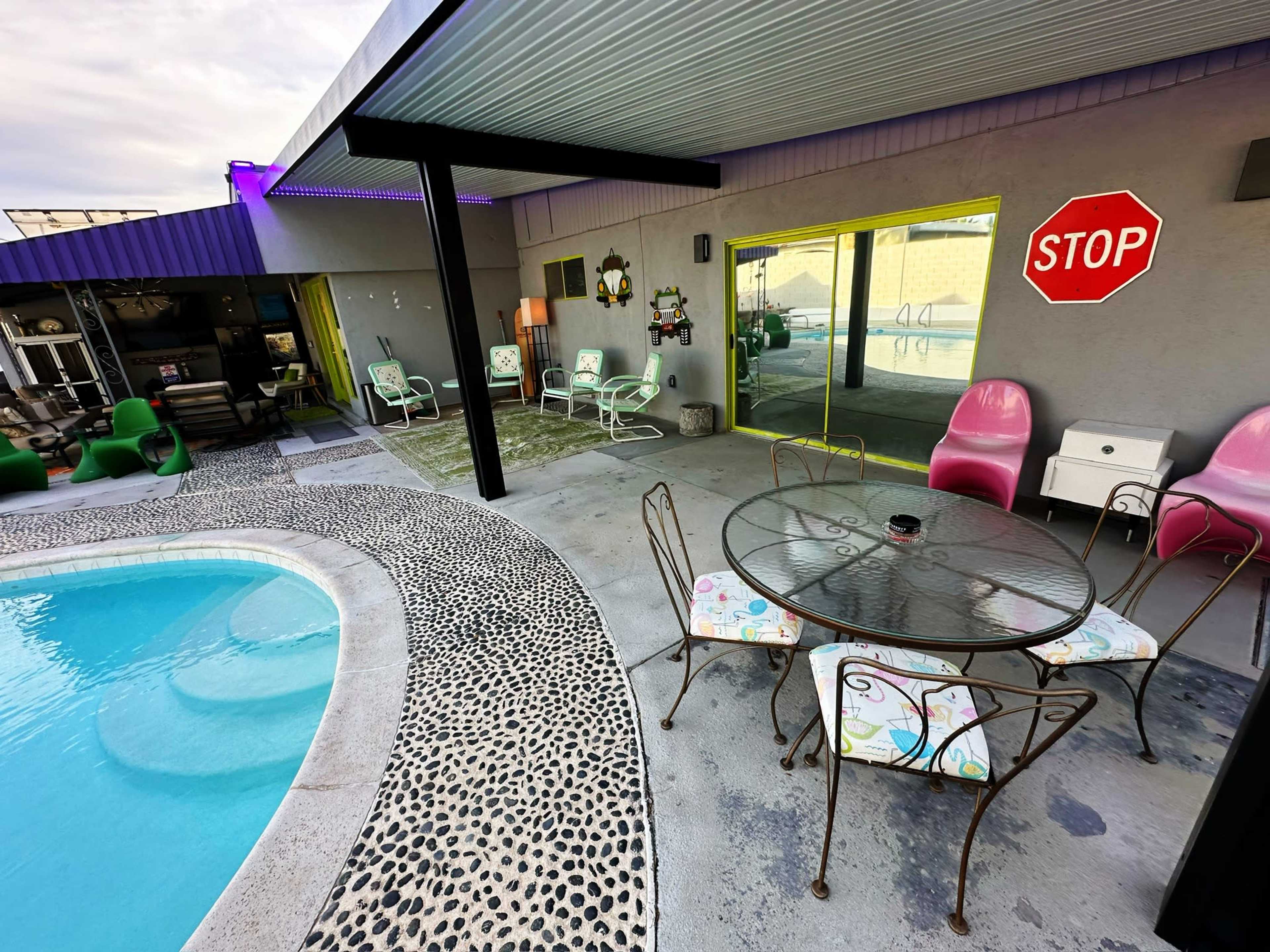 A poolside area features a round glass table with floral chairs, green and pink patio furniture, and a "STOP" sign on the wall.