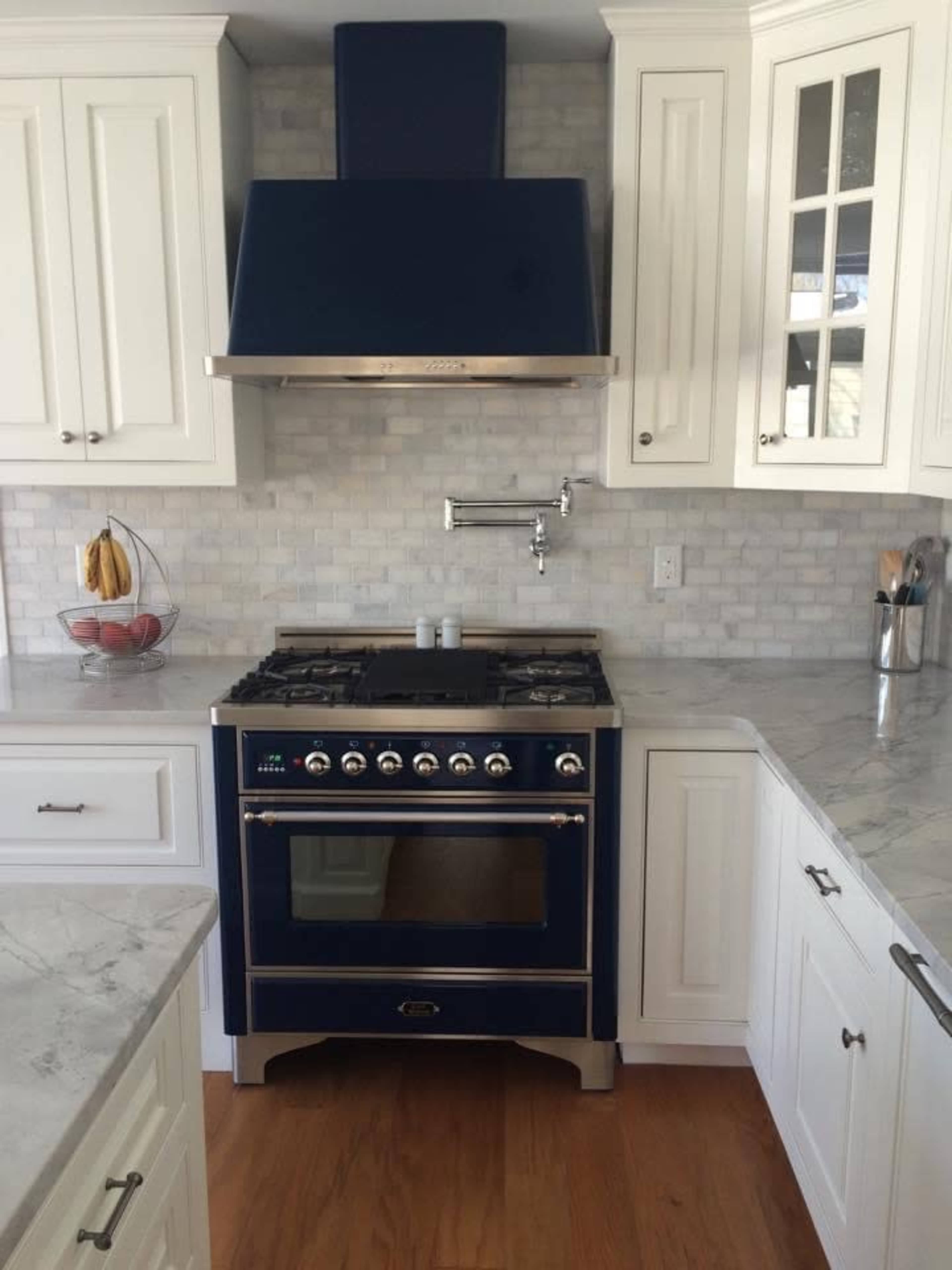 A modern kitchen featuring a blue gas range with a stainless steel hood, white cabinetry, a marble countertop, and a tiled backsplash.