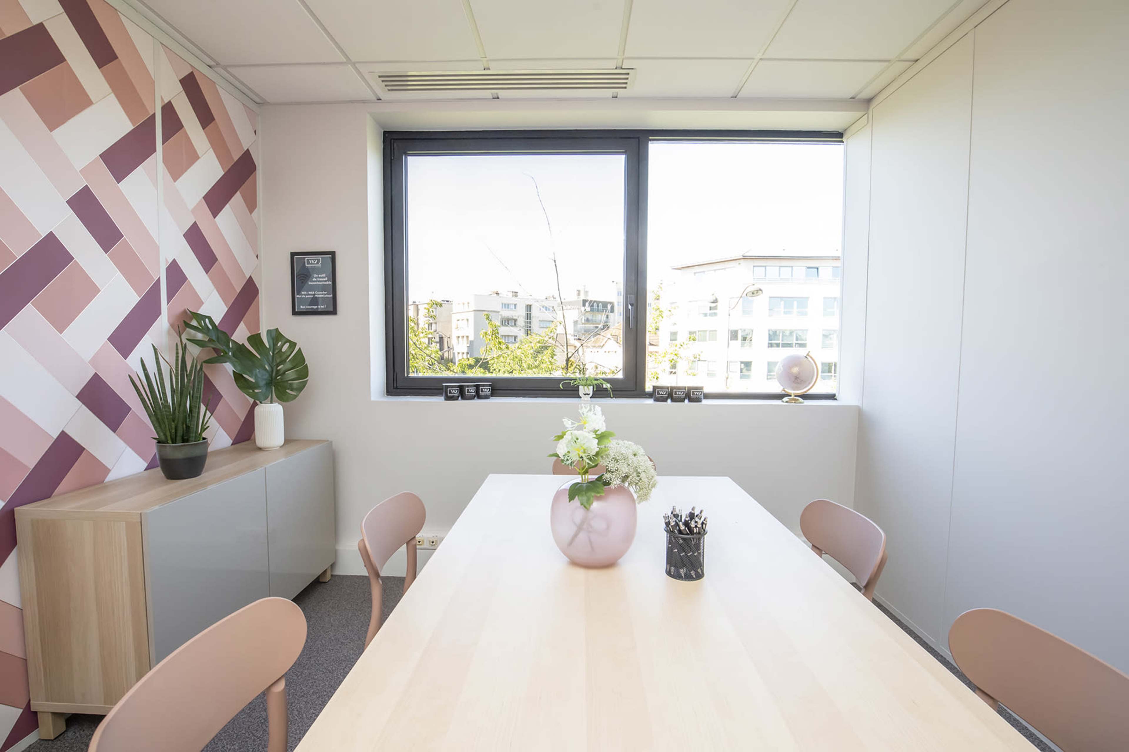 A bright office meeting room features a wooden table surrounded by minimalist chairs, a window overlooking a city view, and a decorative plant and flowers on the table.