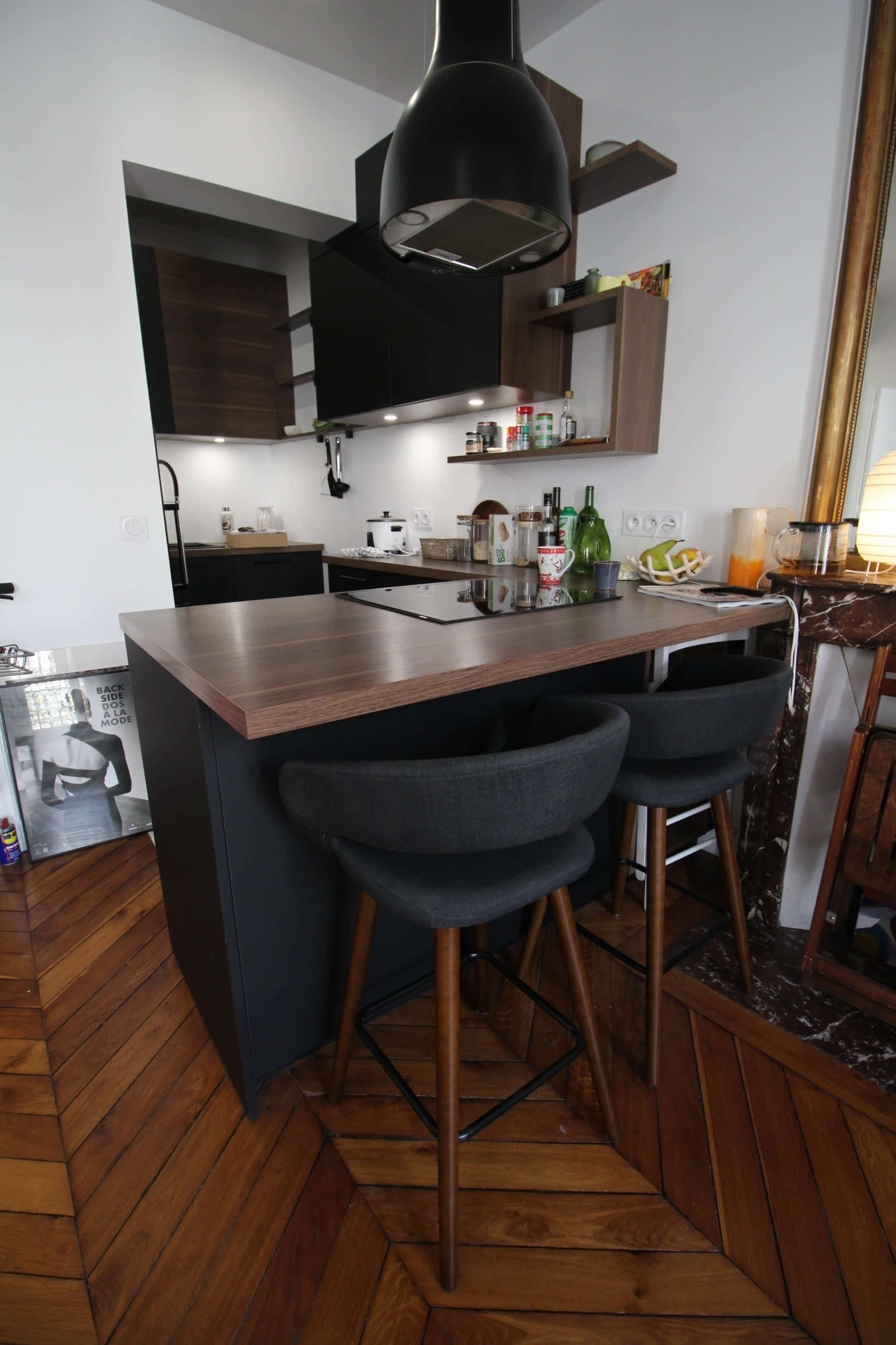 The image shows a modern kitchen with a wooden bar counter and two black upholstered stools, featuring a minimalist design and sleek cabinetry.