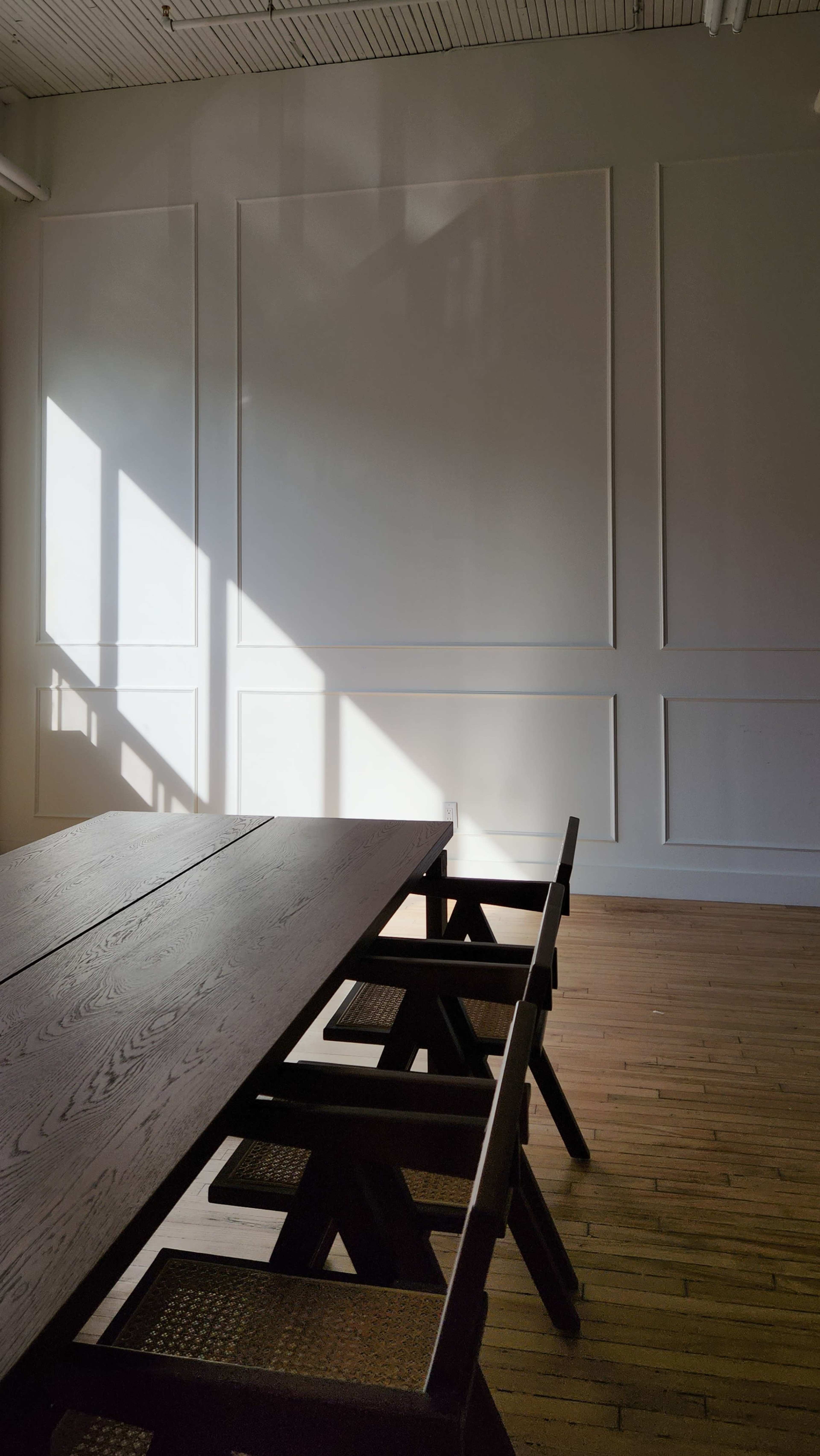 A long wooden table with chairs is positioned in a brightly lit room, featuring a shadow cast on the white wall.