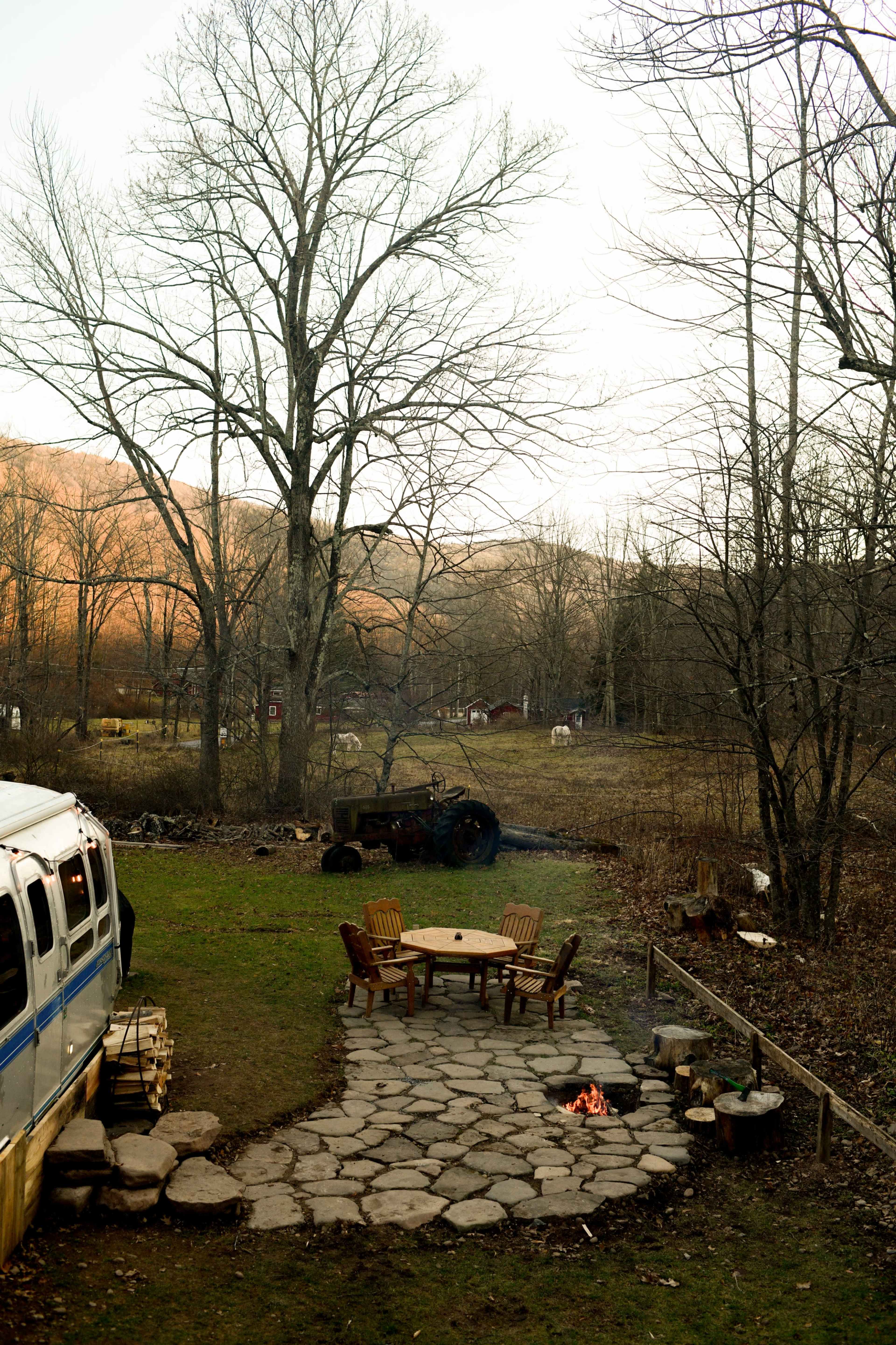 A stone patio with chairs surrounds a small fire pit, and an old truck is parked in a rural field with trees and a van in the foreground.