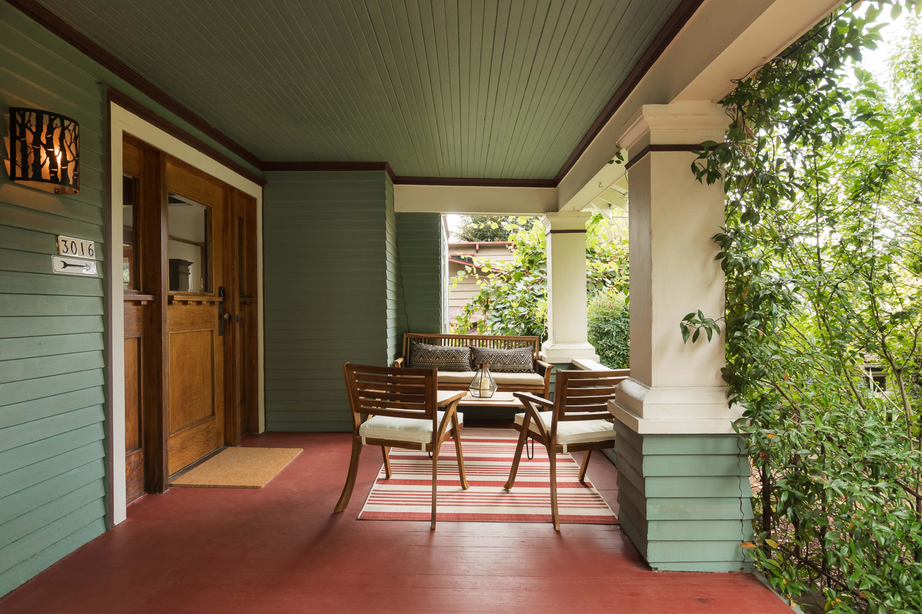 A porch features a seating area with two chairs and a small table, surrounded by greenery and detailed with a patterned rug.