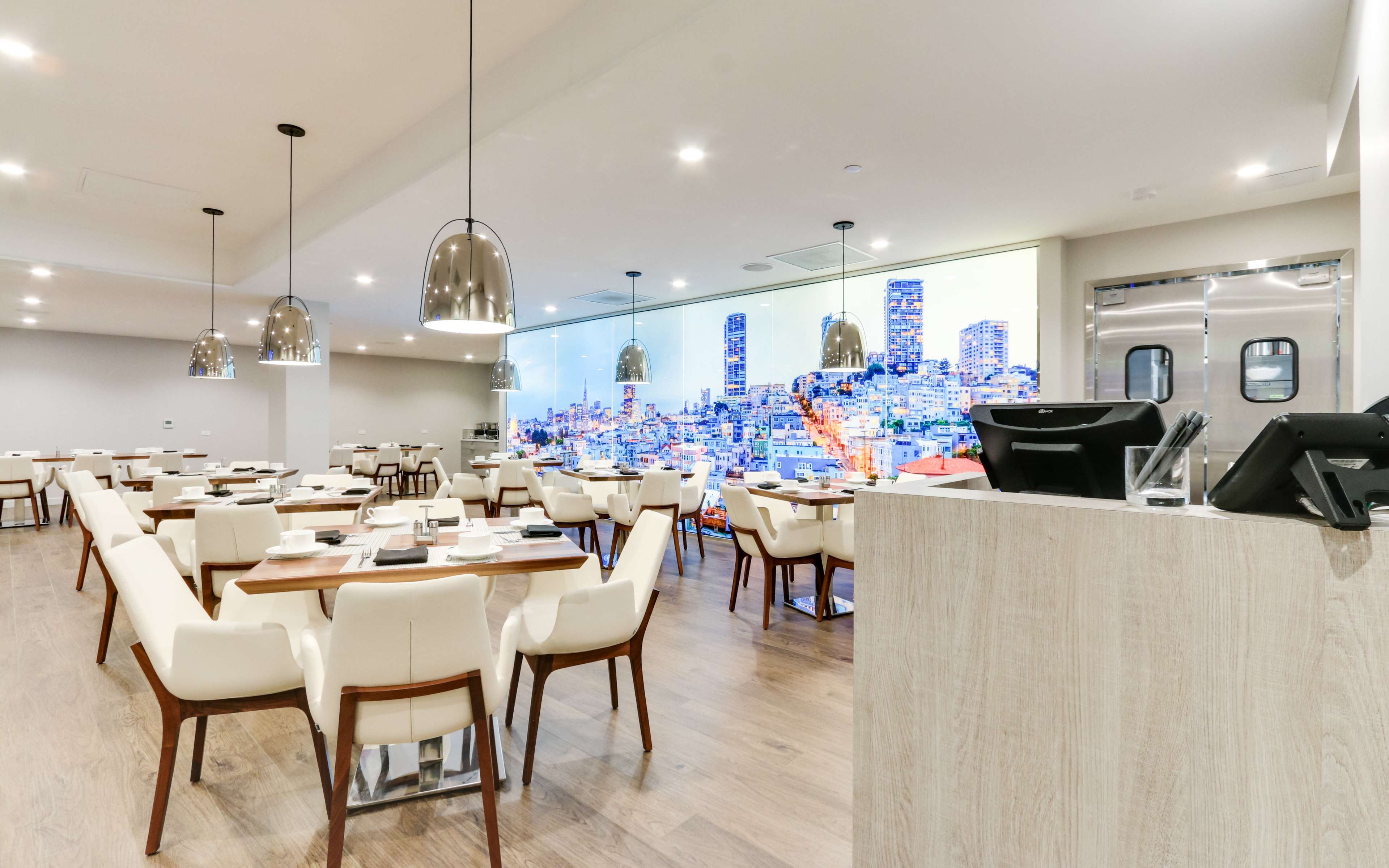 The image shows a modern restaurant interior with wooden flooring, white tables, and chairs, illuminated by pendant lights, featuring a large digital cityscape mural on one wall.