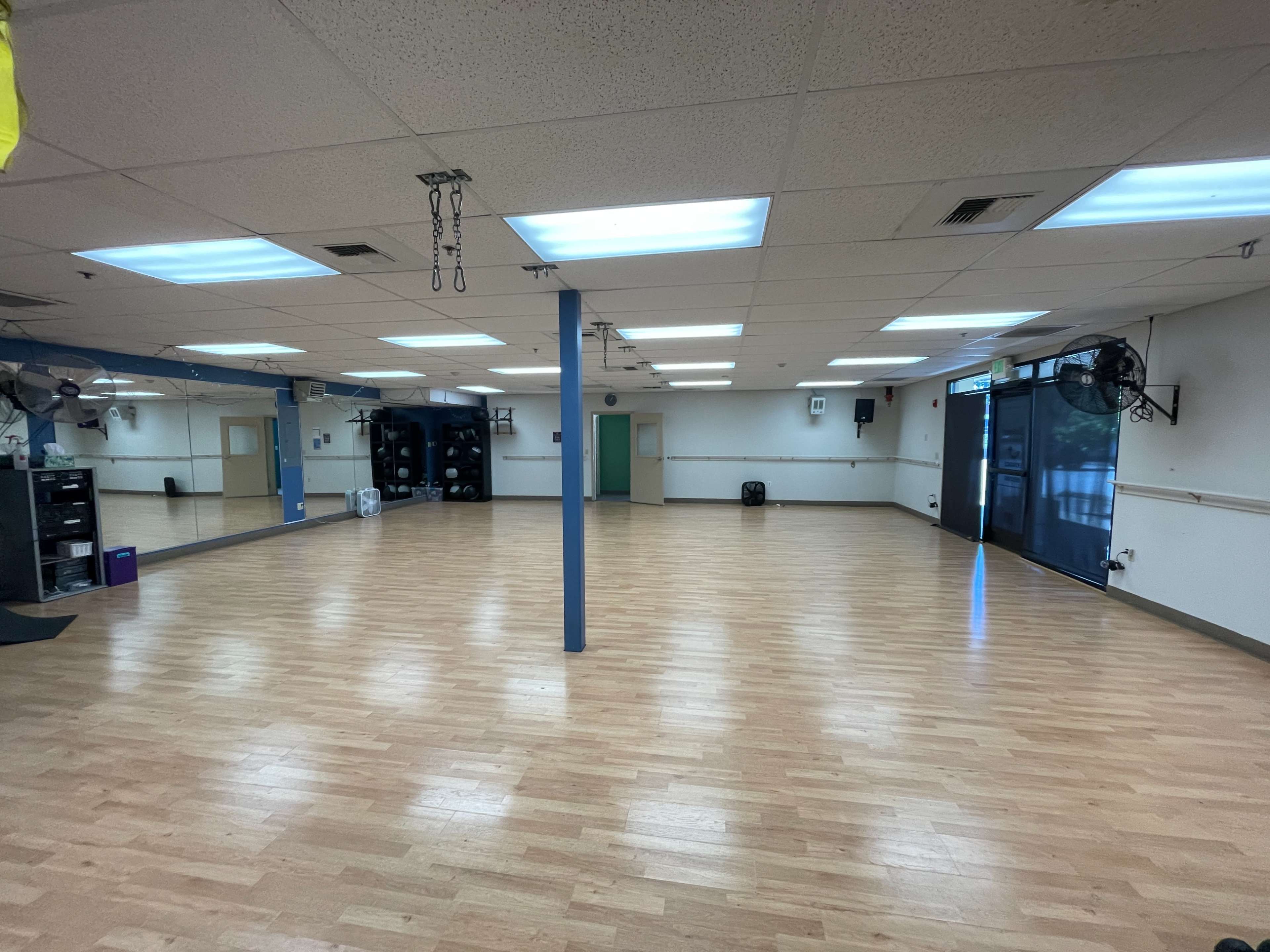 A spacious, empty room with wooden flooring, fluorescent lights, and large mirrors on one wall.