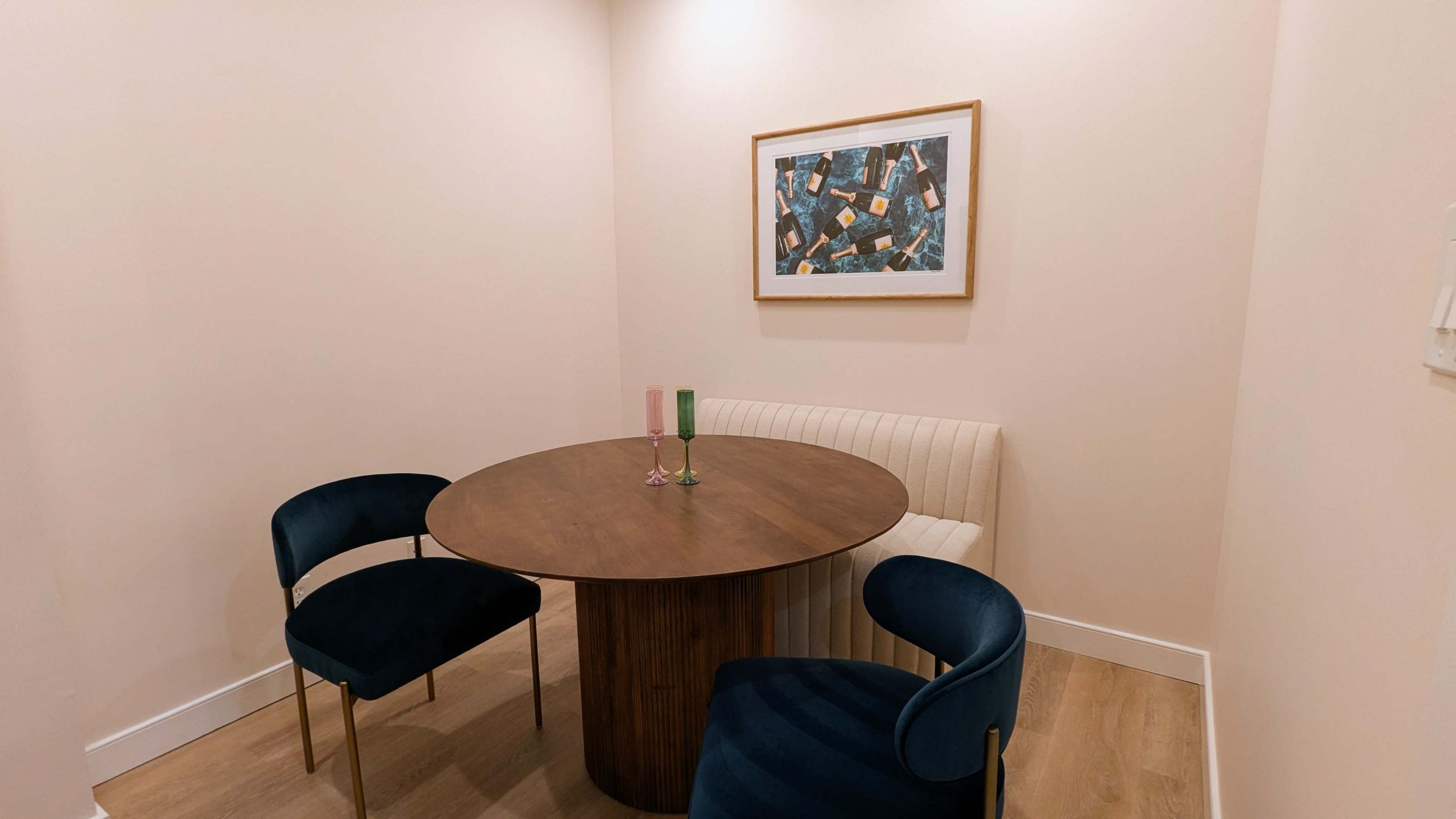 A round wooden table is set with two glass candle holders in a small dining area with beige walls and a framed abstract artwork.