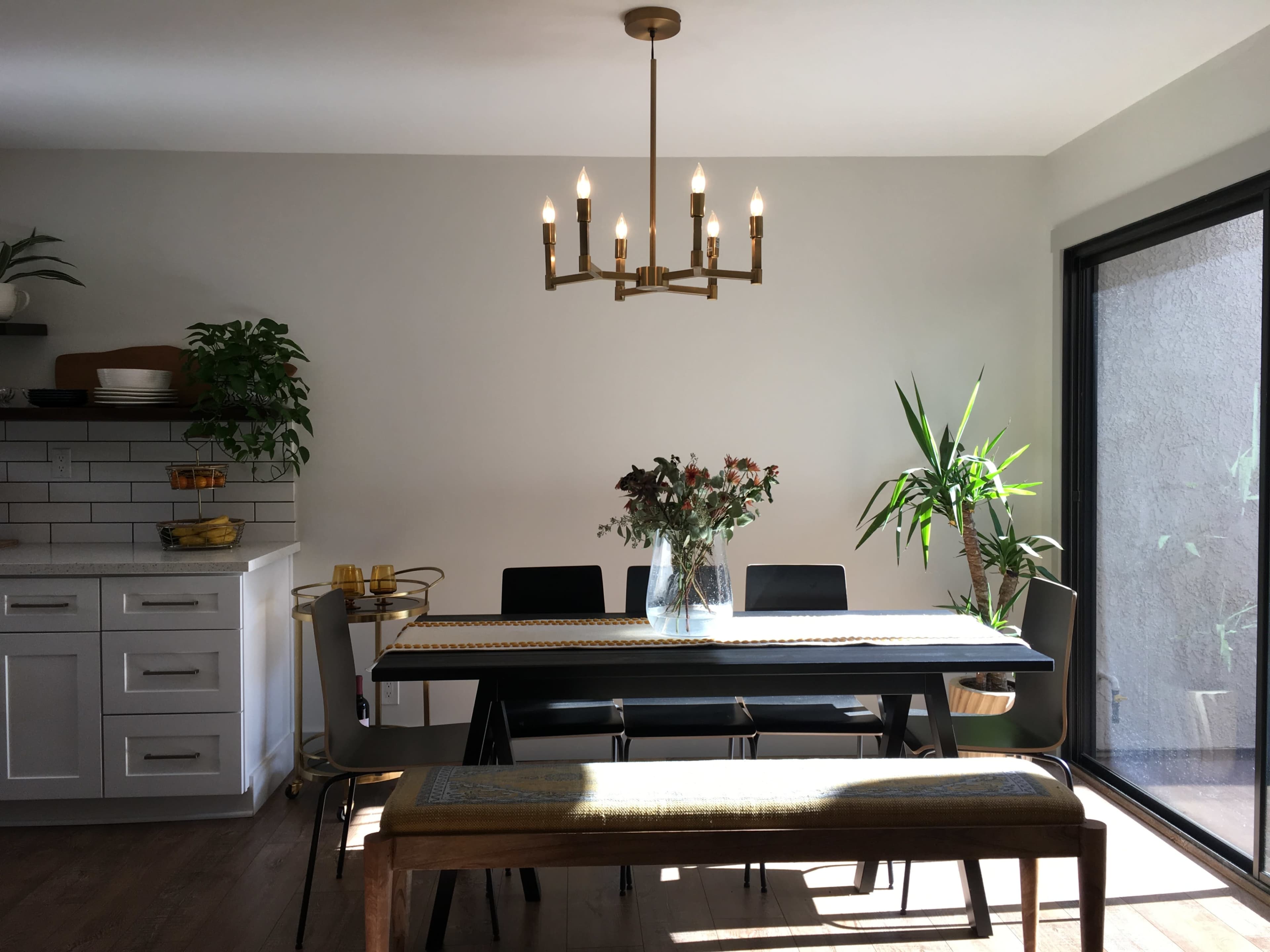 A modern dining area featuring a long table with chairs, a central chandelier, and plants by the window.