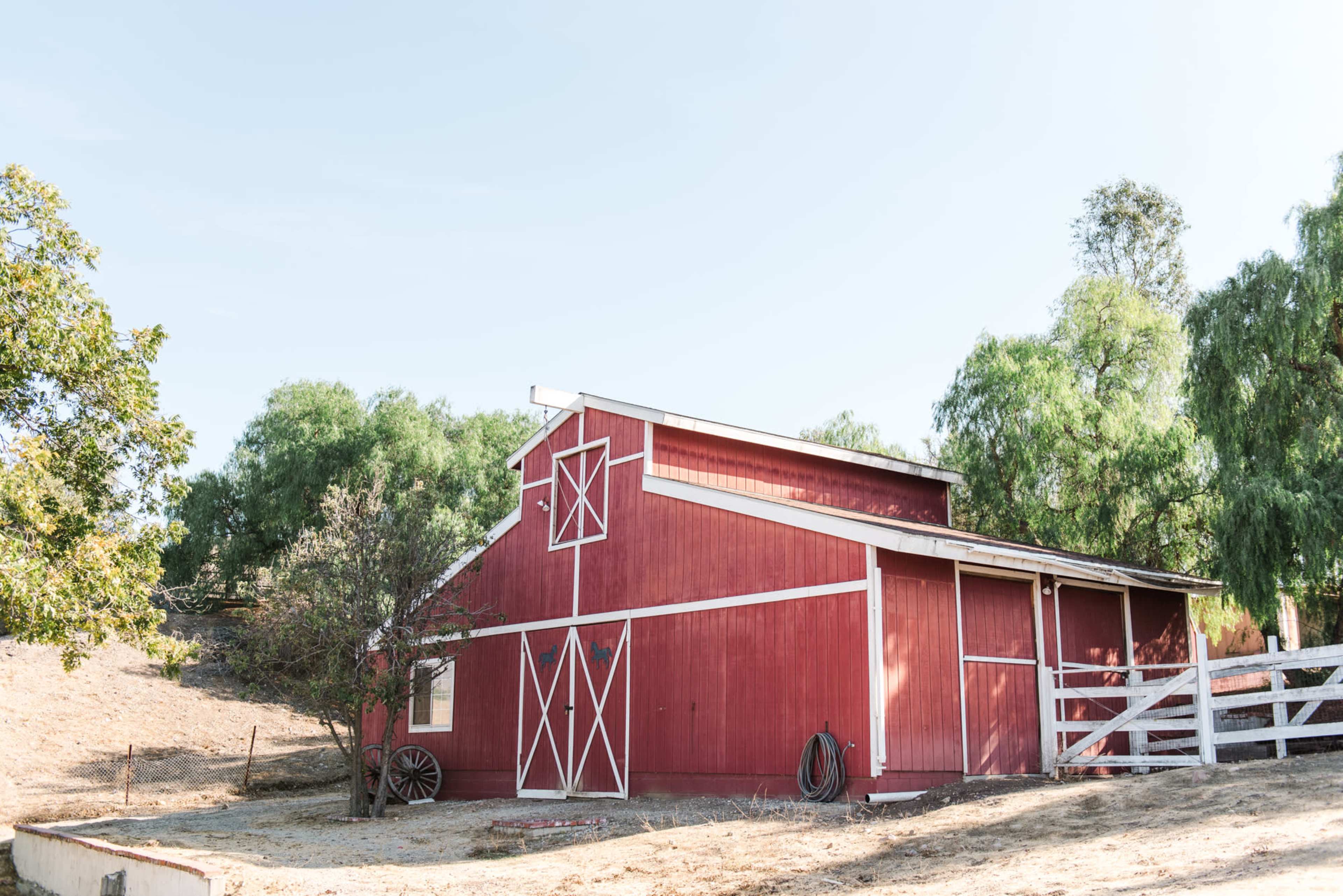 A red barn with white trim is positioned on a dry hillside surrounded by various trees.