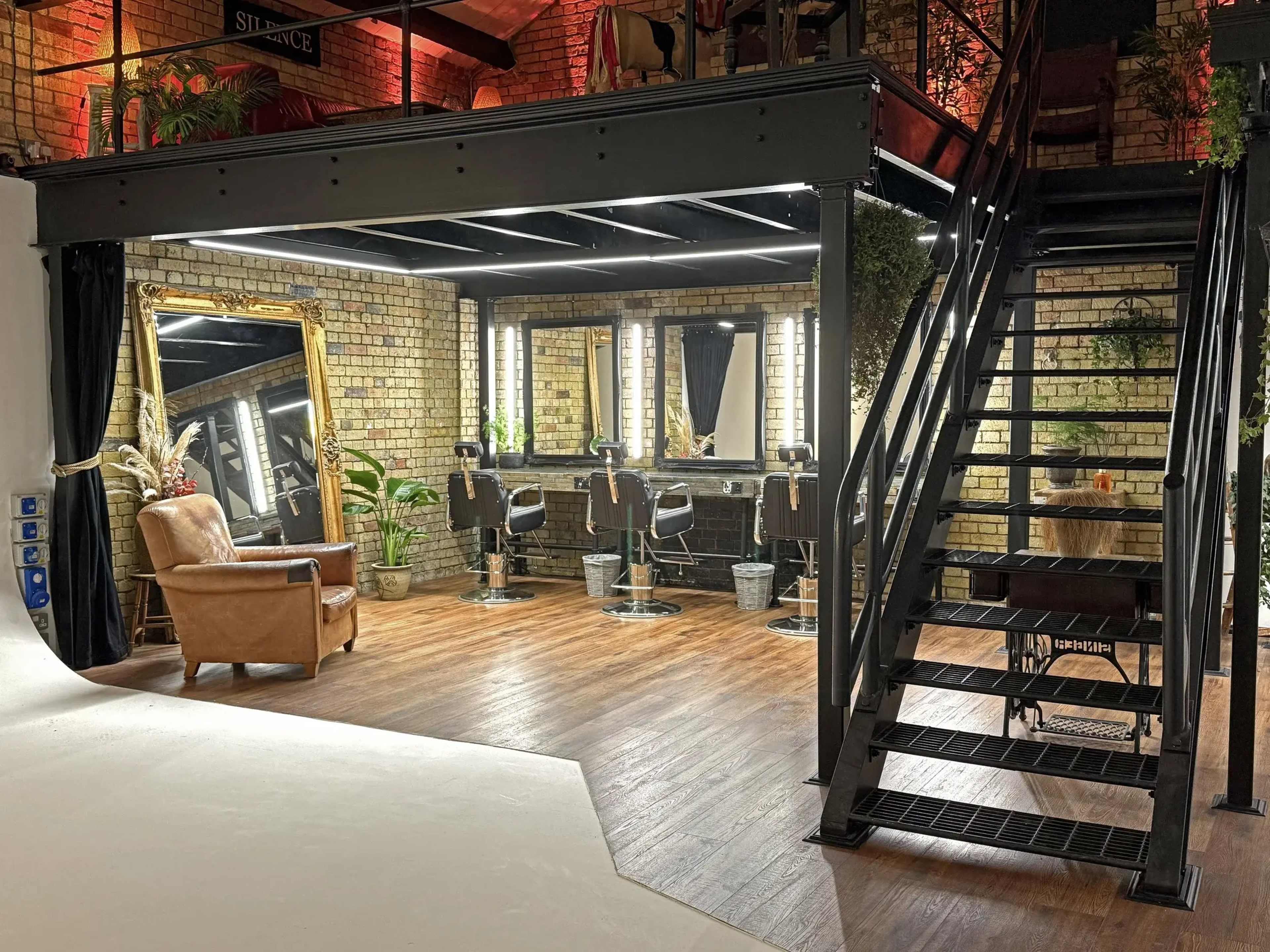 The image shows a stylist salon featuring a black metal staircase, a leather armchair, several styling chairs with mirrors, and brick walls adorned with plants.