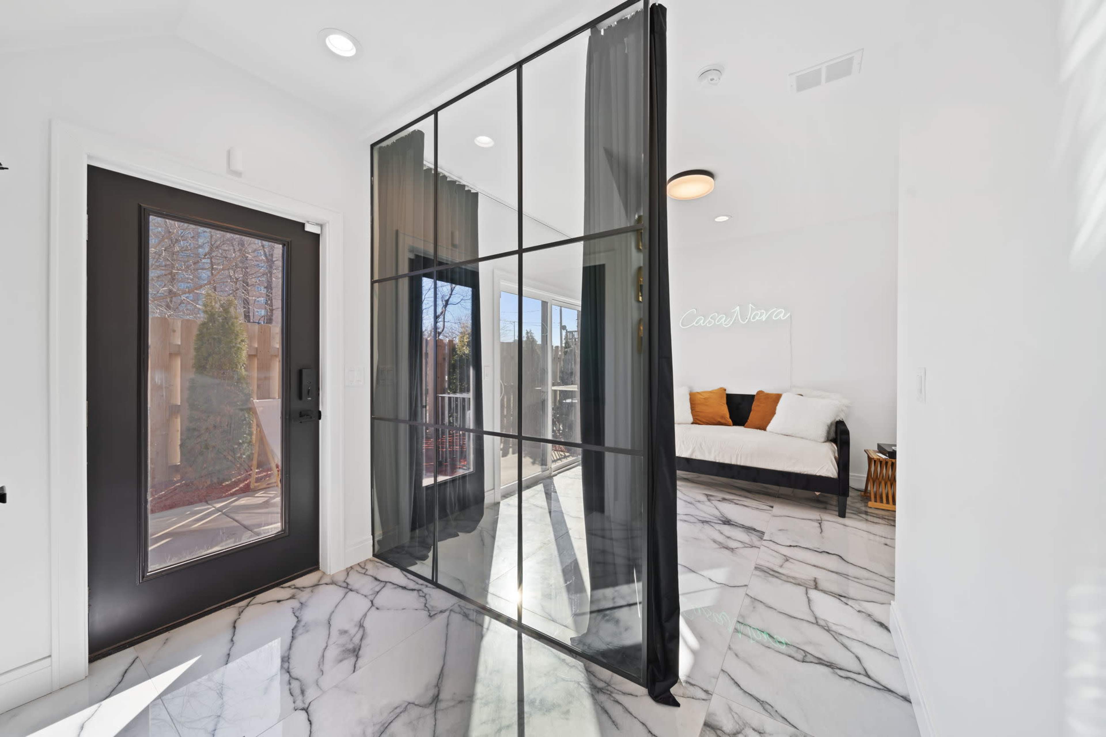 The image shows a modern entryway featuring a large black-framed glass partition, a white sofa with orange and brown cushions, and marble flooring.