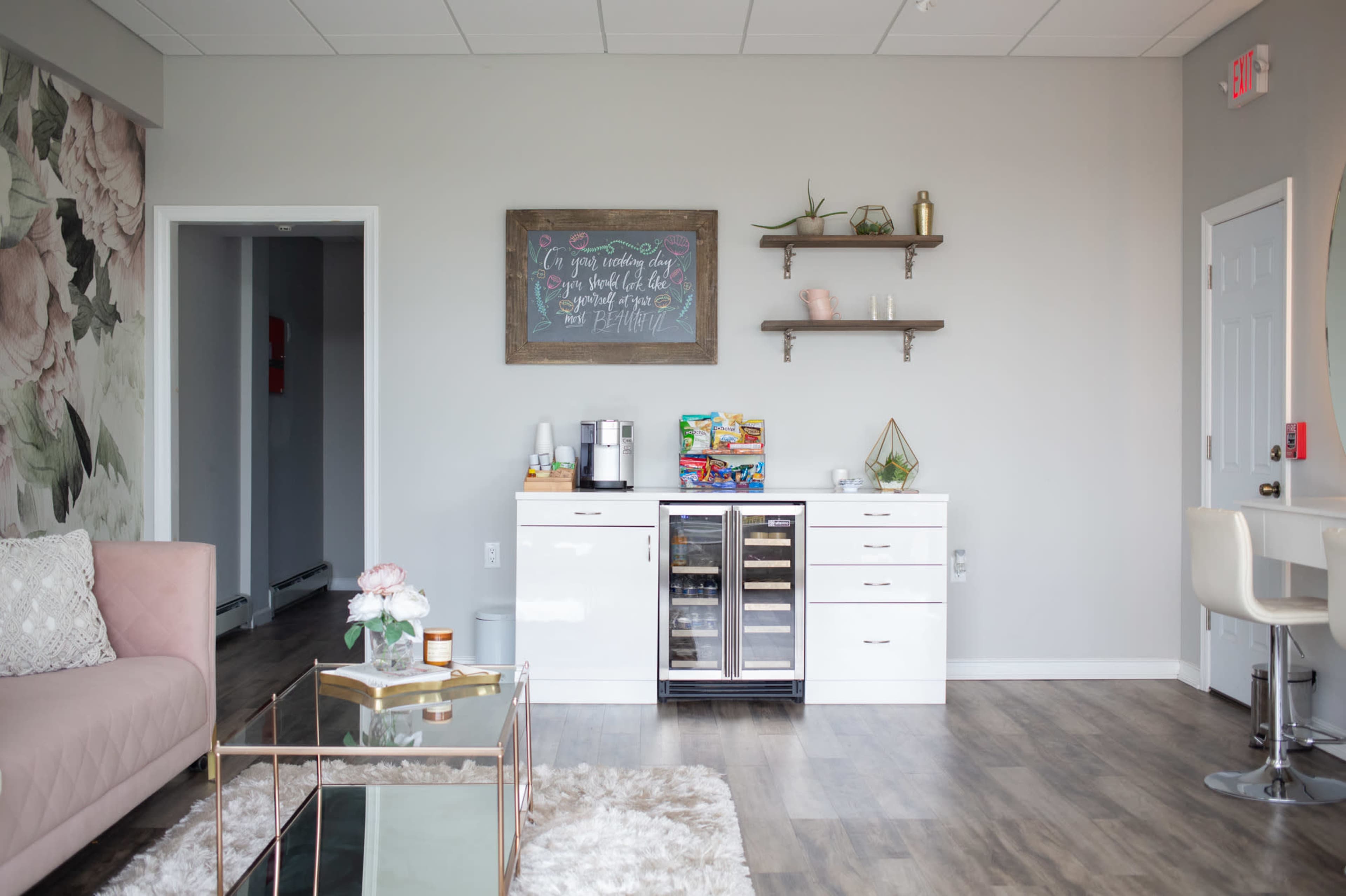 A modern waiting area with a pink sofa, a coffee machine, a beverage refrigerator, and decorative shelves against a light gray wall.