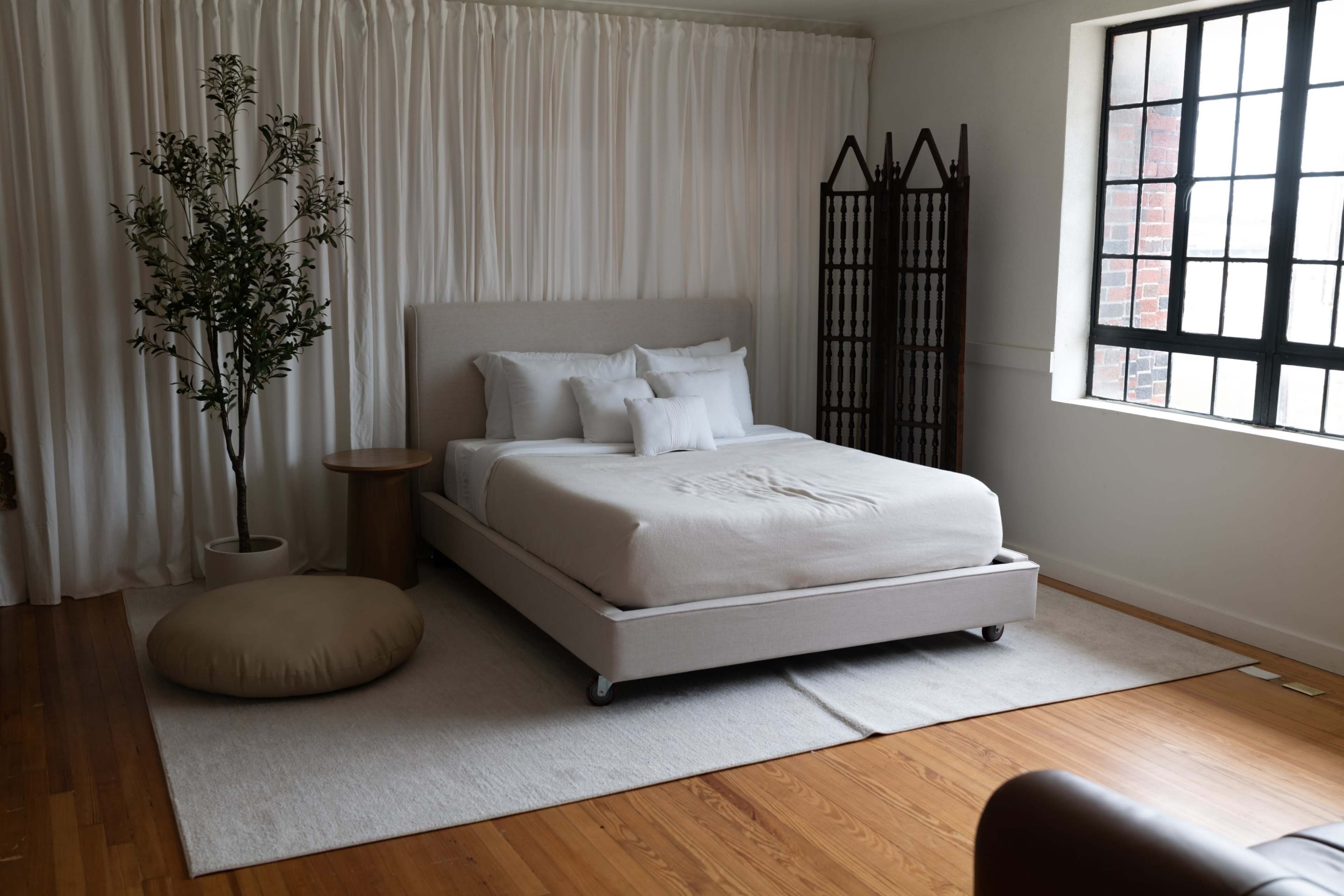 A minimalist bedroom featuring a bed, a potted plant, a round ottoman, and a folding screen against a light wall with large windows.