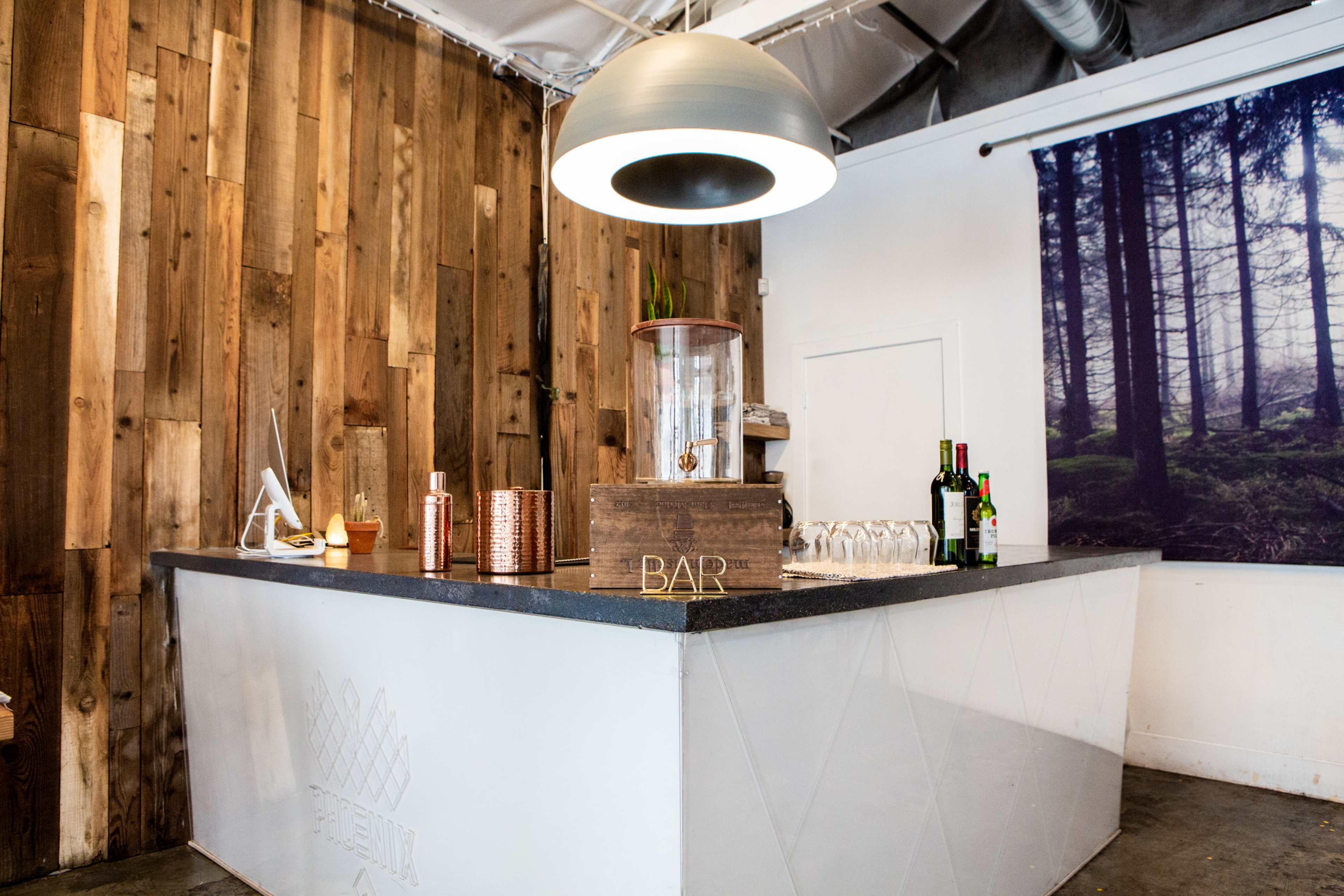 The image shows a modern bar counter made of wood and concrete, featuring a large pendant light and a selection of bottles and glasses neatly arranged on the surface.