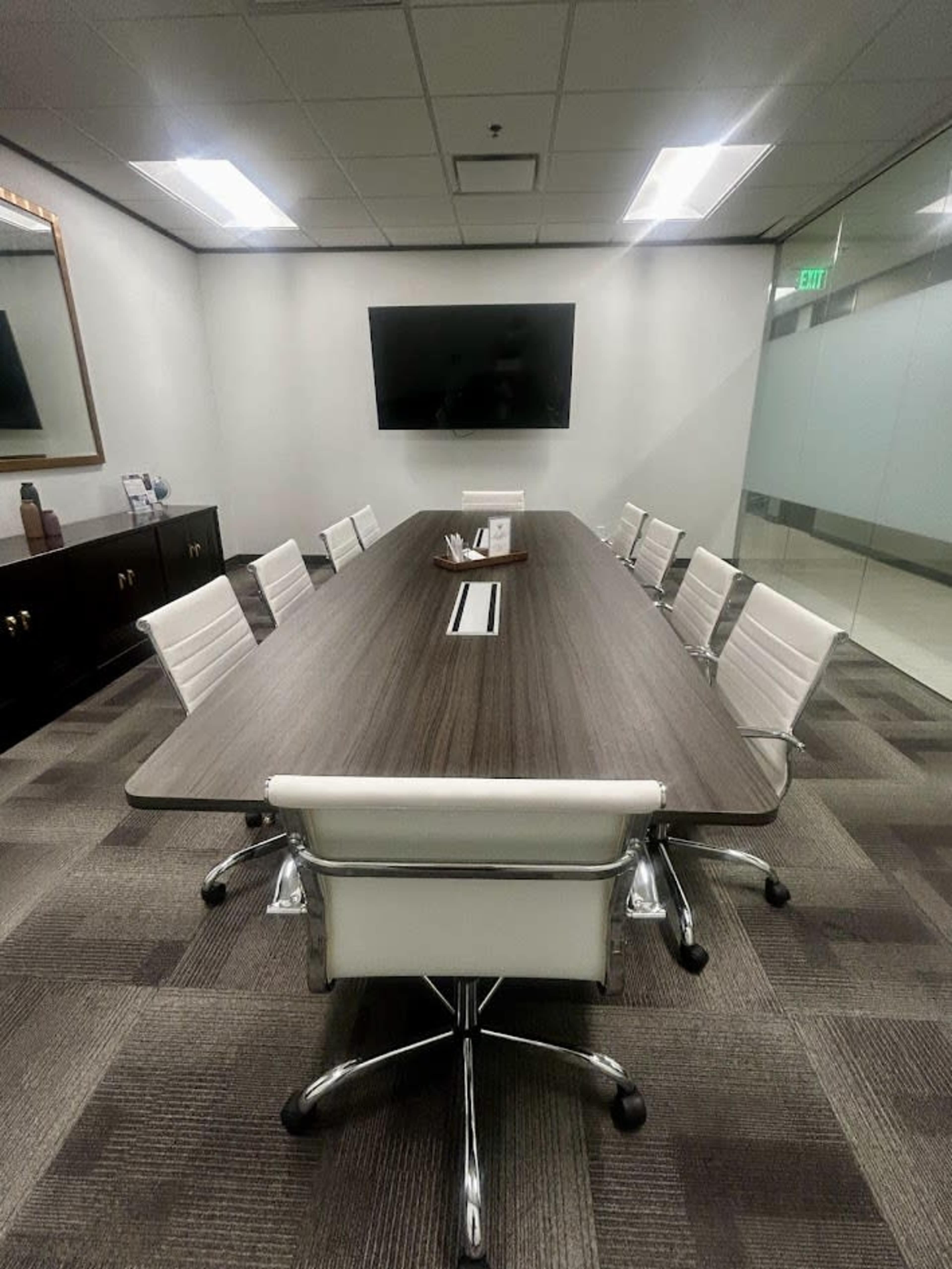 The image shows a modern conference room featuring a long wooden table surrounded by white leather chairs, with a television and a large mirror on the walls.