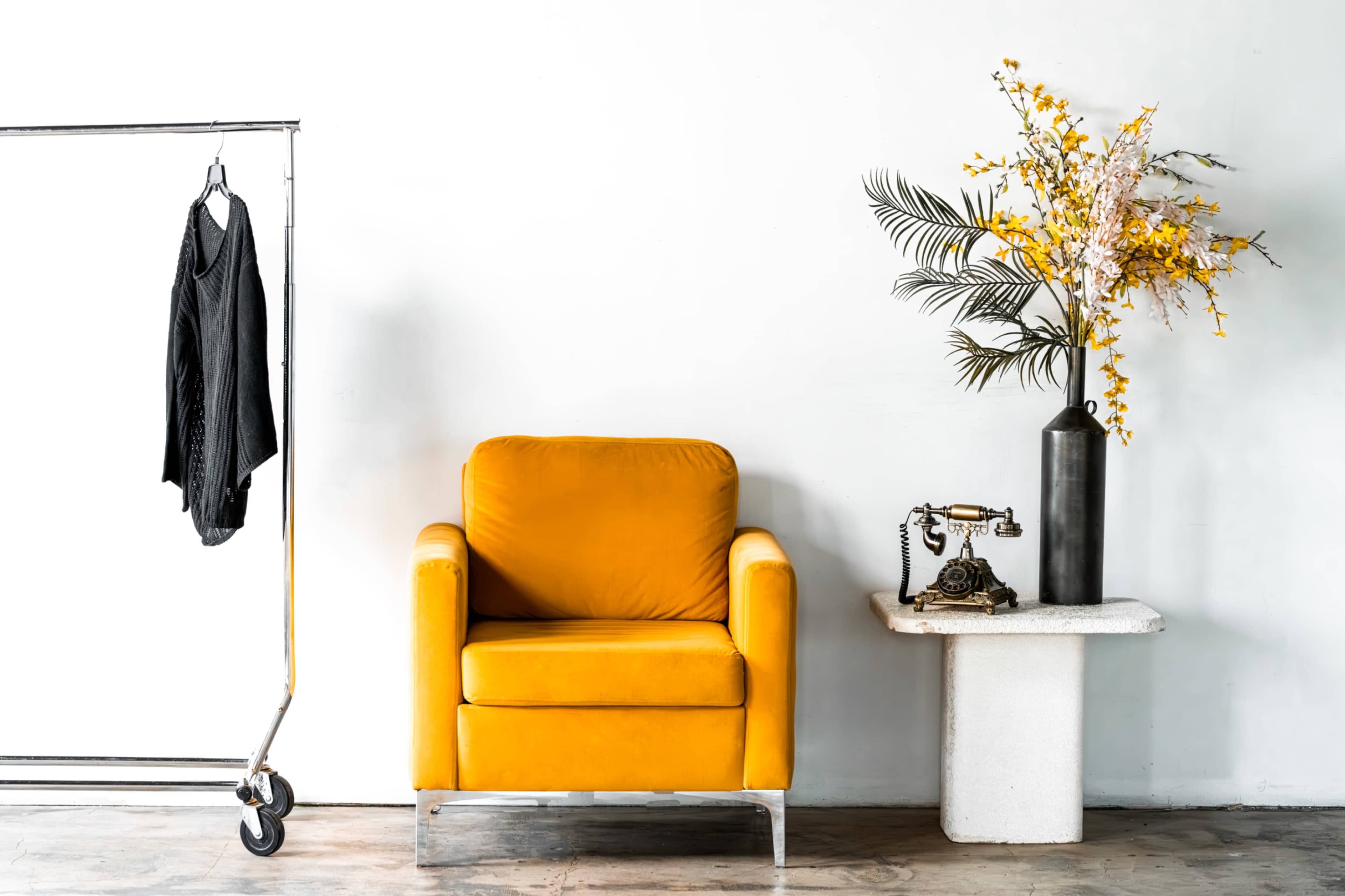 A yellow upholstered armchair is positioned next to a white concrete side table, which holds a decorative vase with flowers and an antique telephone, while a clothing rack displays a black garment.