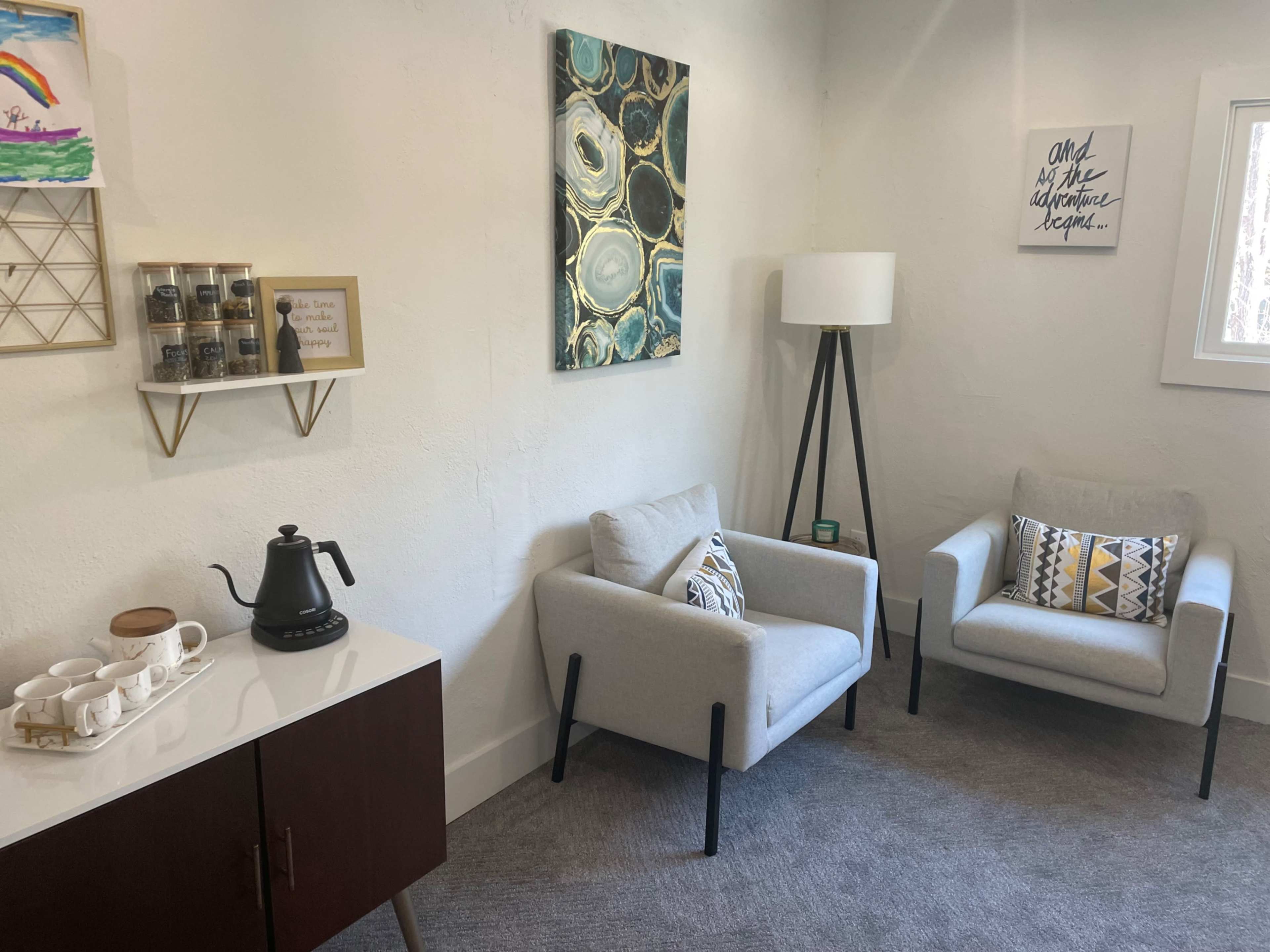 The image depicts a modern, cozy waiting area featuring two light-colored chairs, a coffee station with a kettle and tea supplies, and decorative wall art.
