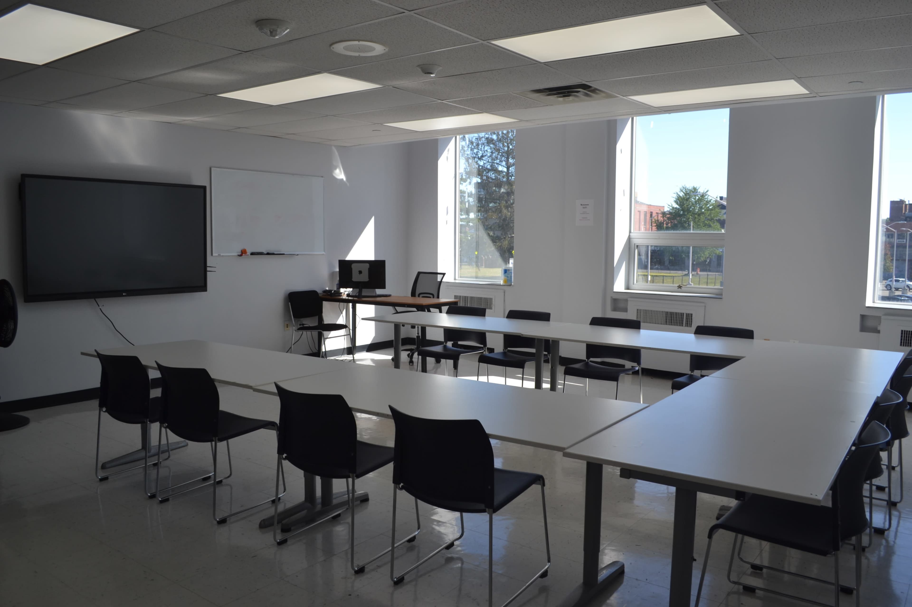 The image shows a classroom with several tables arranged in a U-shape, black chairs, a large screen, and windows allowing natural light.