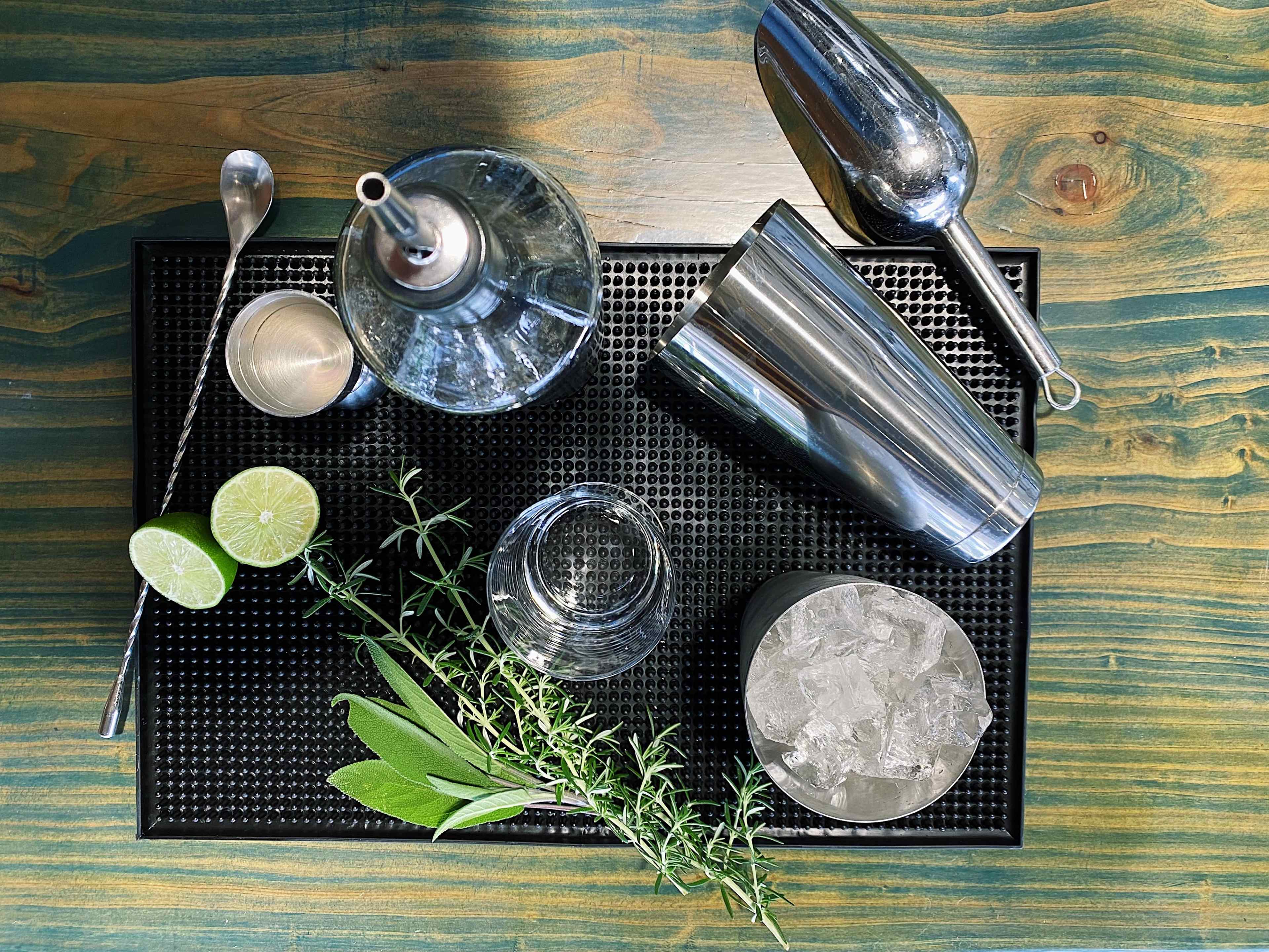 A cocktail preparation setup is arranged on a textured mat, featuring a shaker, mixer, glassware, fresh limes, herbs, and ice.