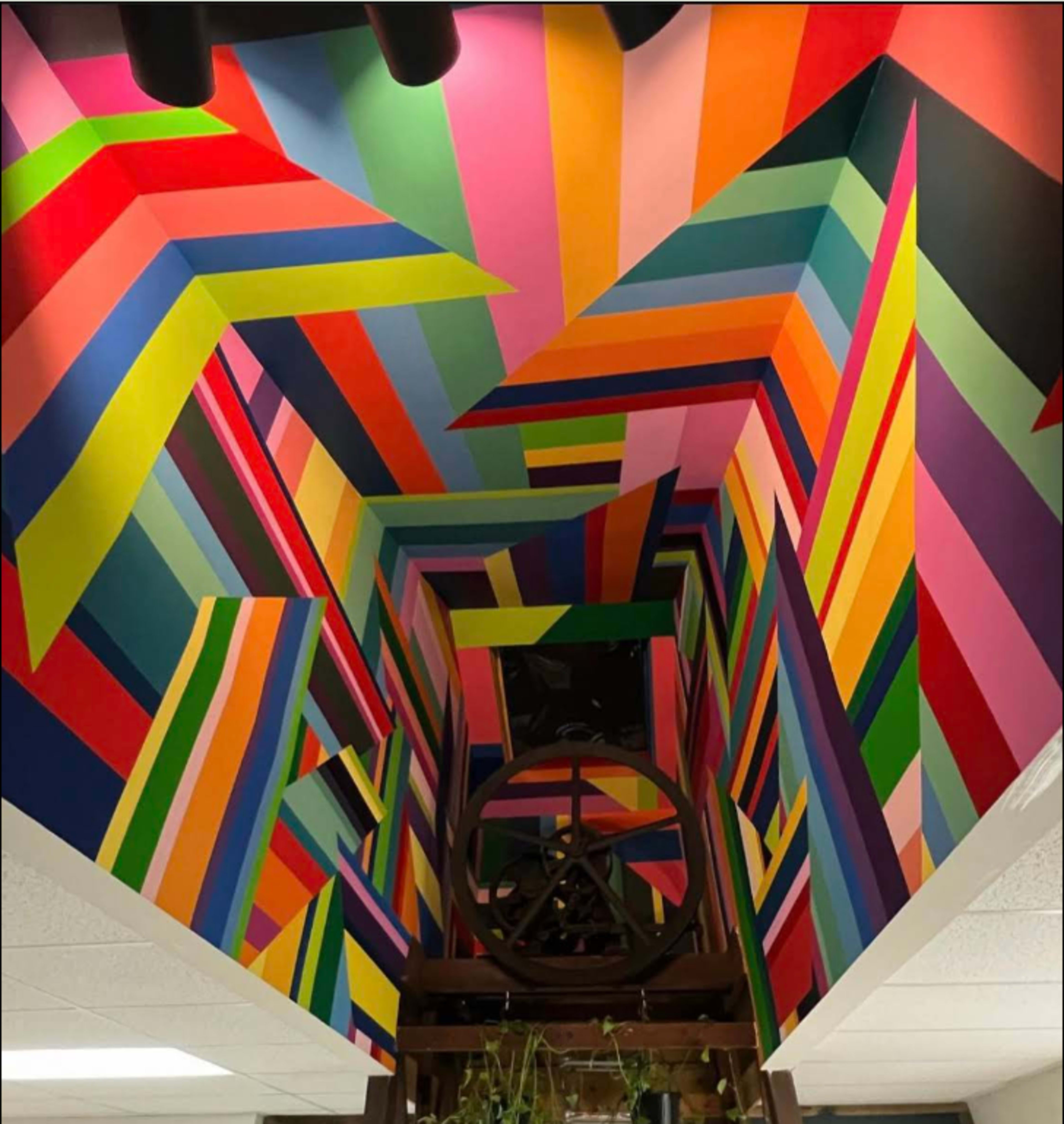 The image features a vibrant, multicolored geometric mural on the ceiling of a stairwell, with sharp angles and contrasting colors.