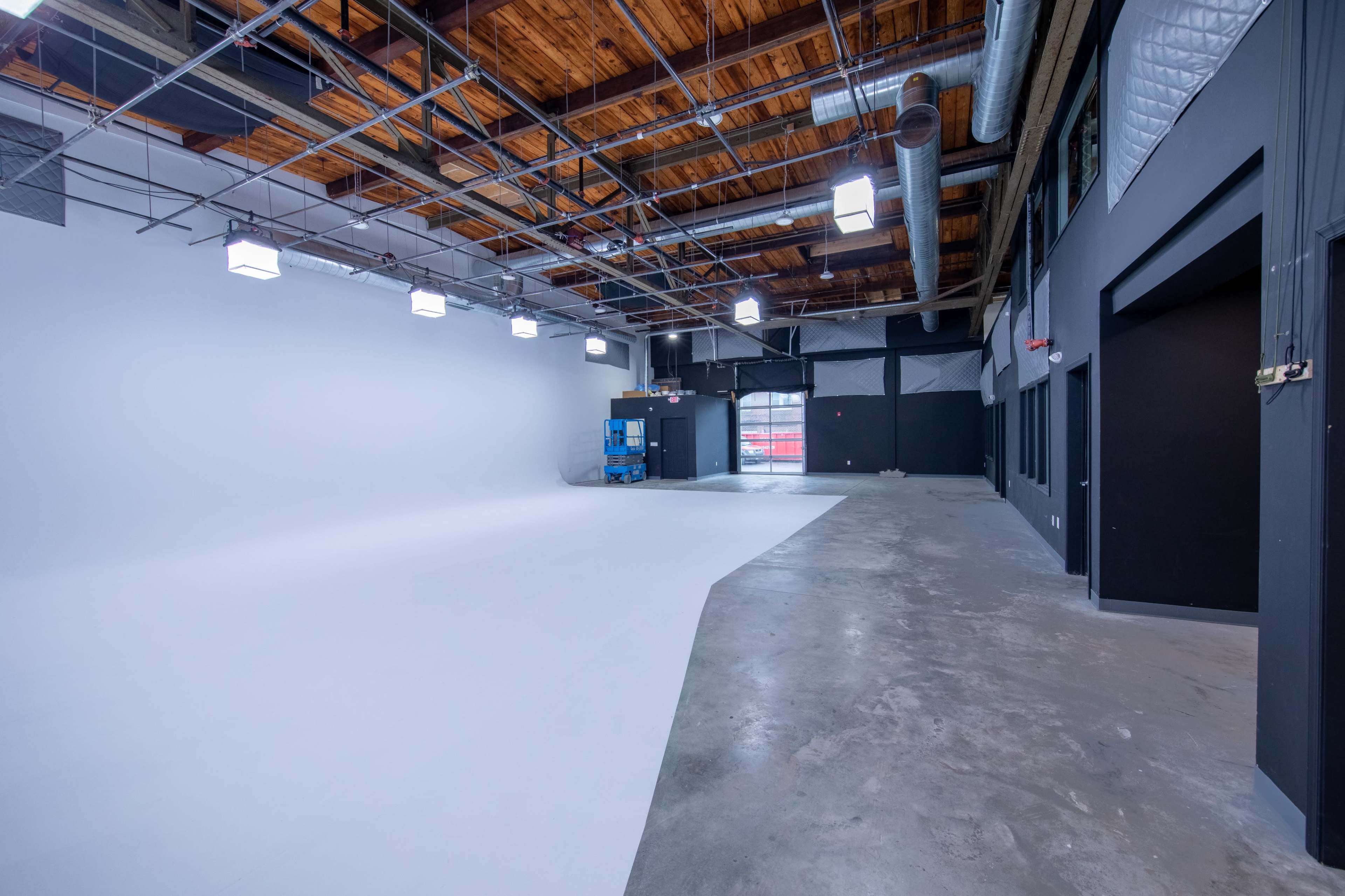 A spacious empty studio with a concrete floor, high wooden beams, and a white backdrop.
