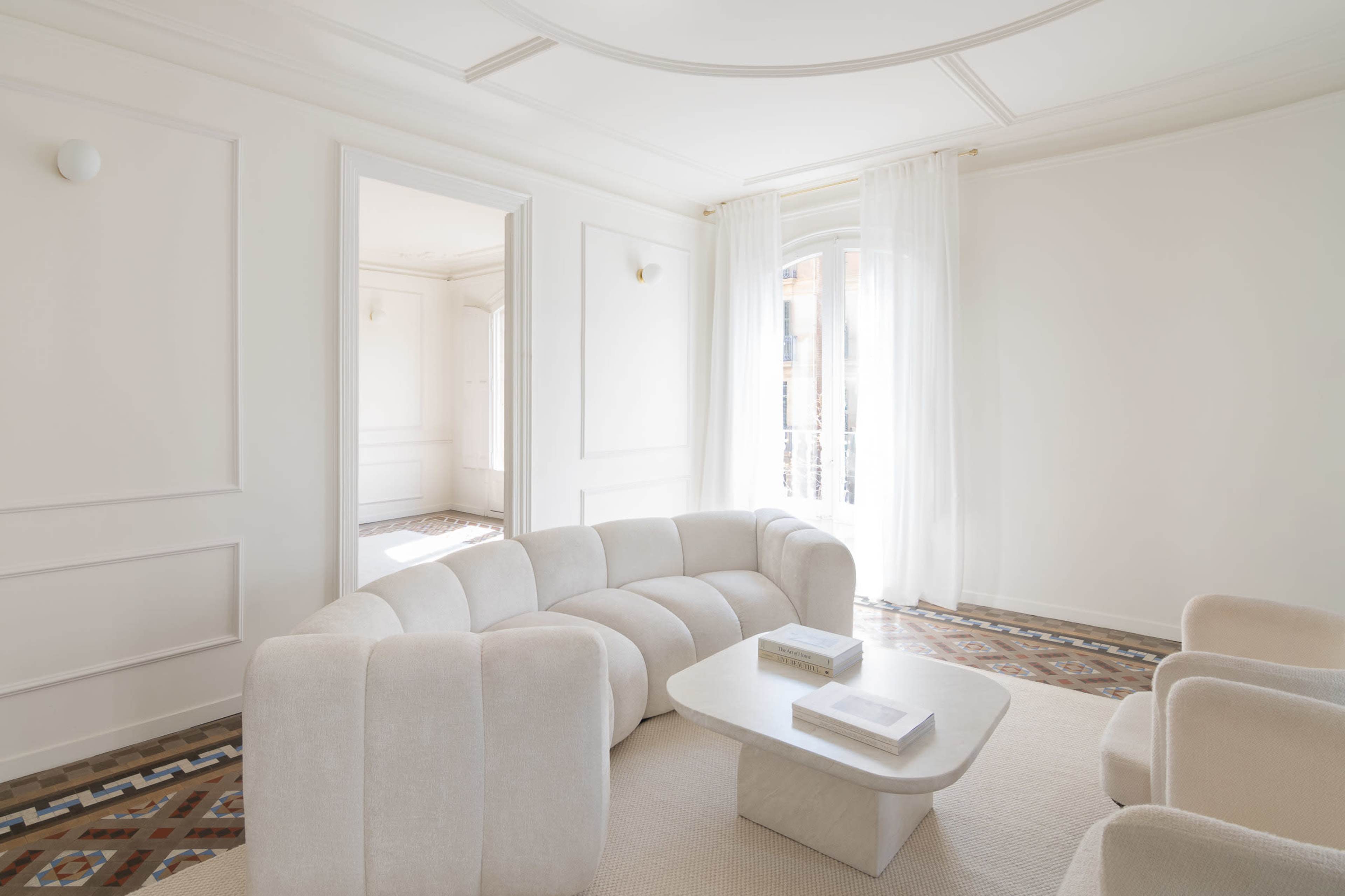 The image shows a bright, minimalist living room featuring a curved sofa, a marble coffee table, and large windows with sheer curtains.