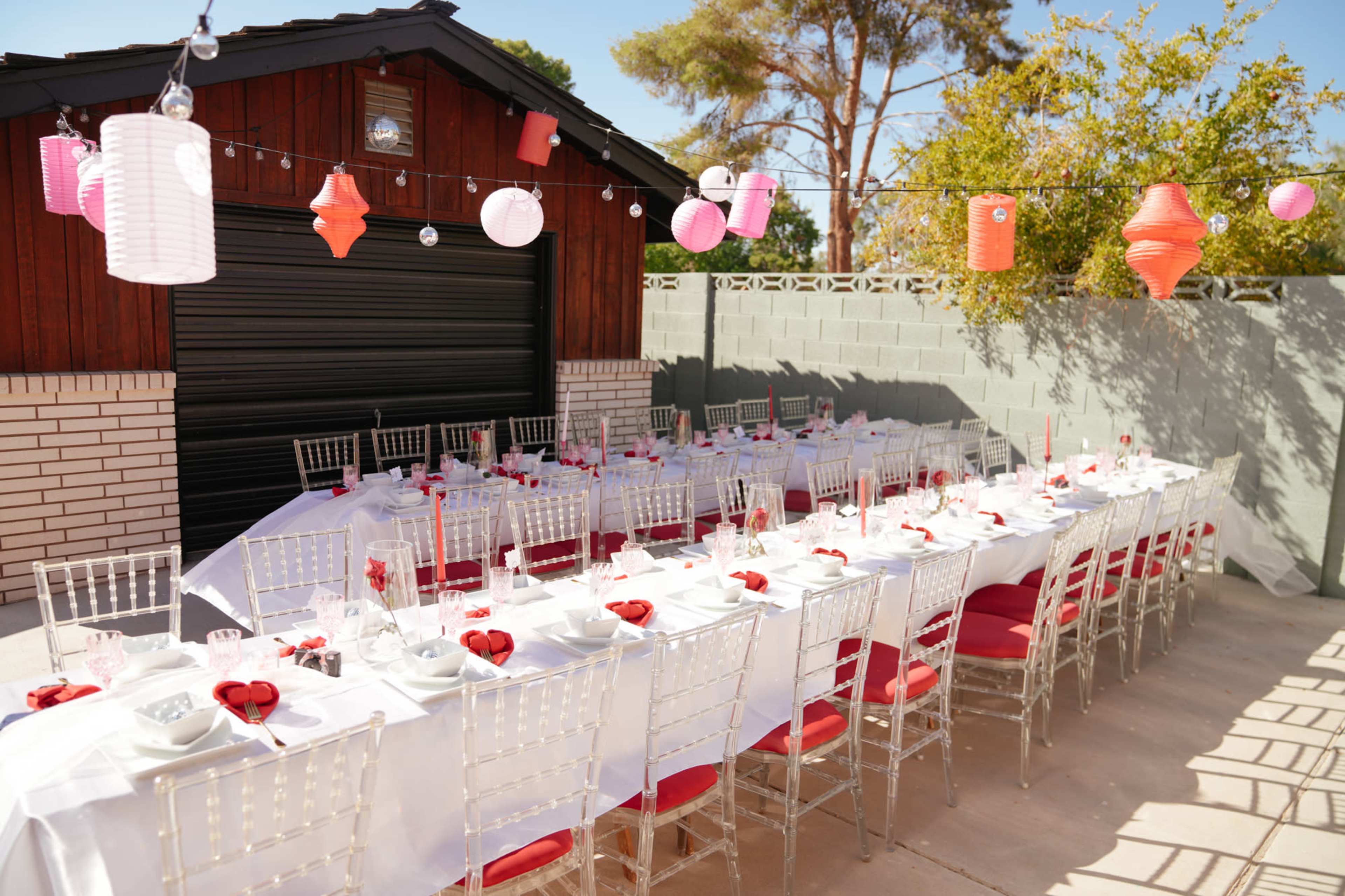 A long table set for a dining event with white tablecloths and red accents, positioned outdoors near a wooden shed decorated with hanging lanterns.