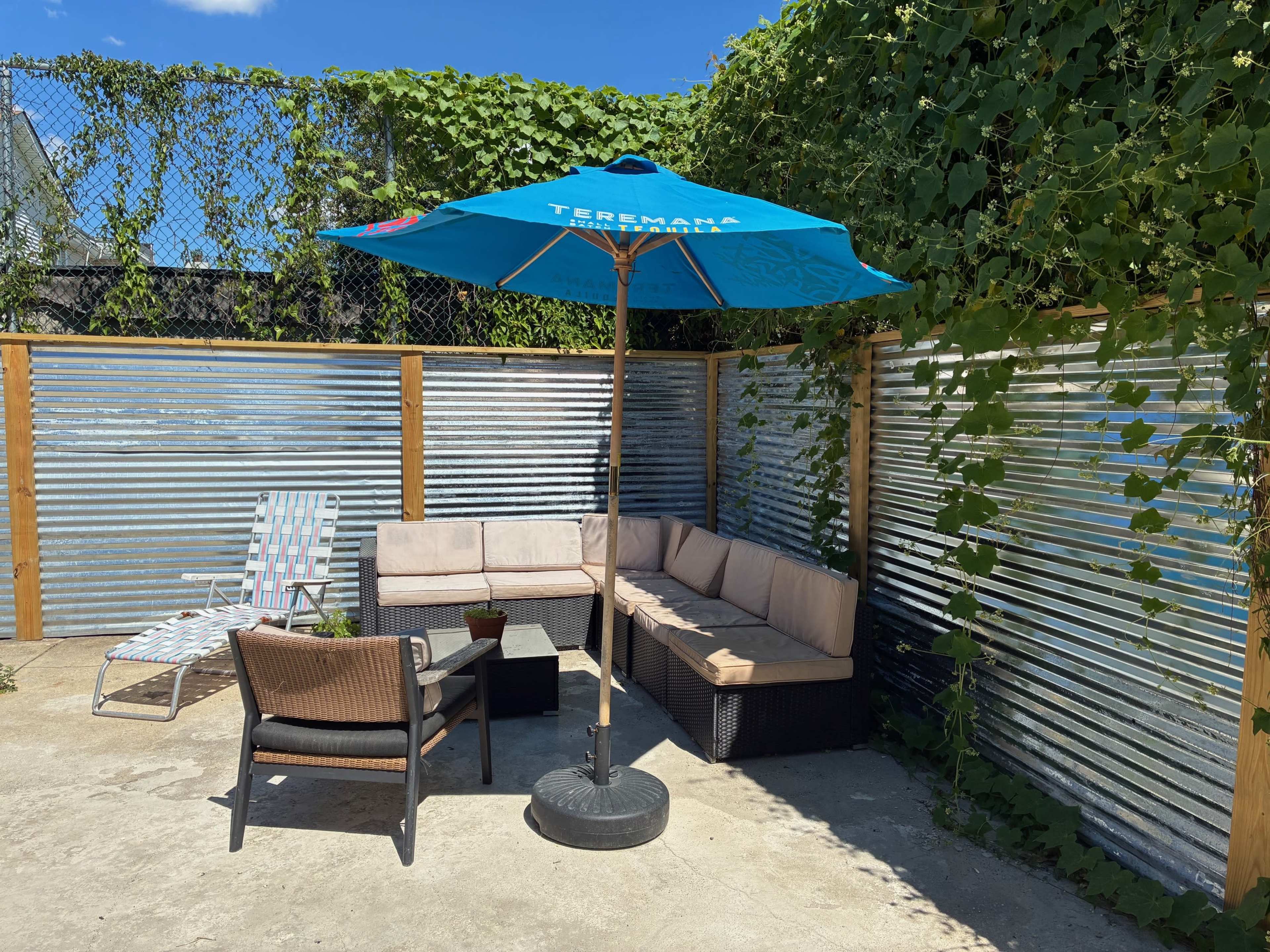 A patio area features a corner seating arrangement with a large blue umbrella, surrounded by metal fencing and greenery.