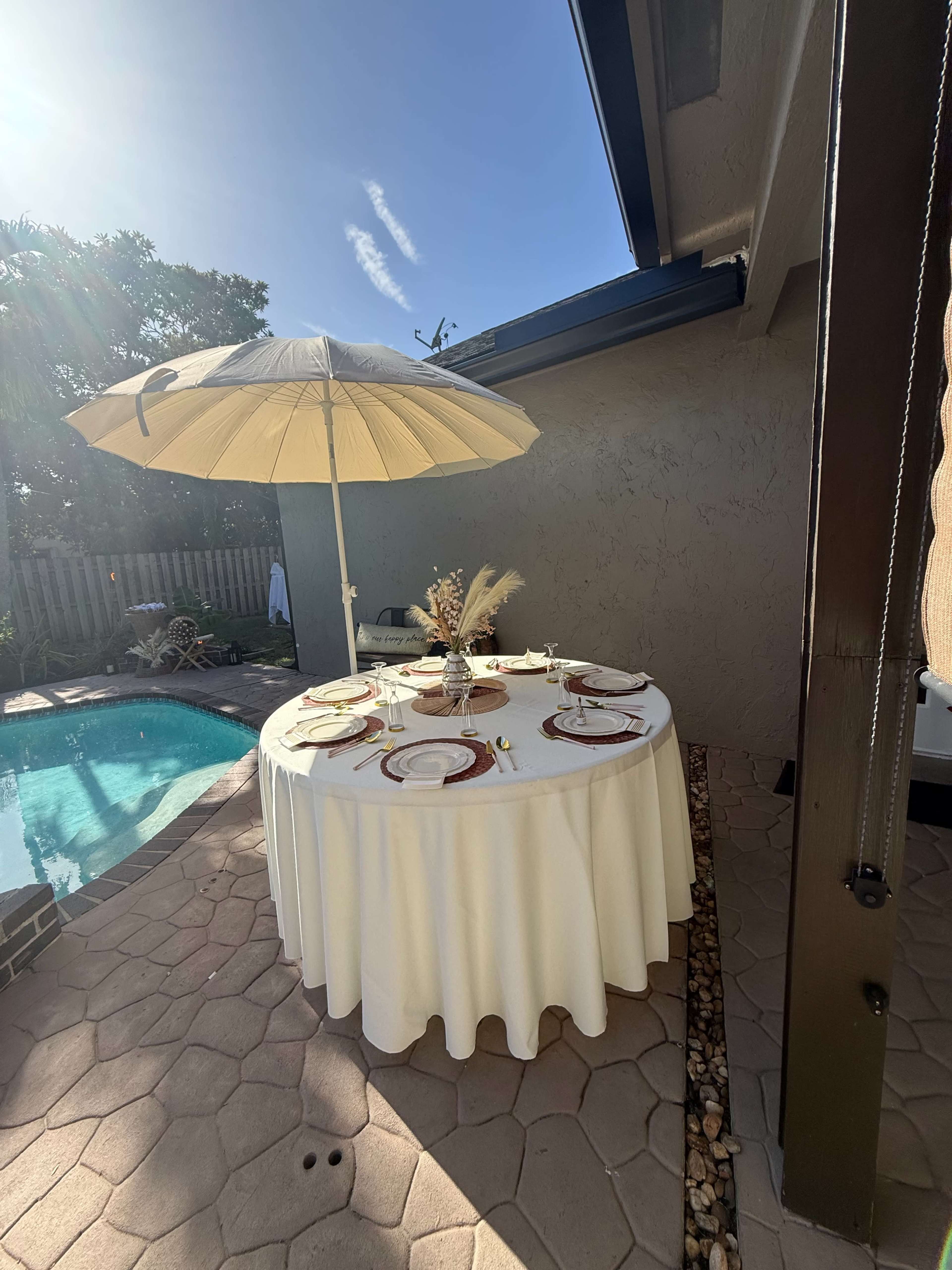 A round dining table with a white tablecloth is set beneath a large umbrella beside a swimming pool.
