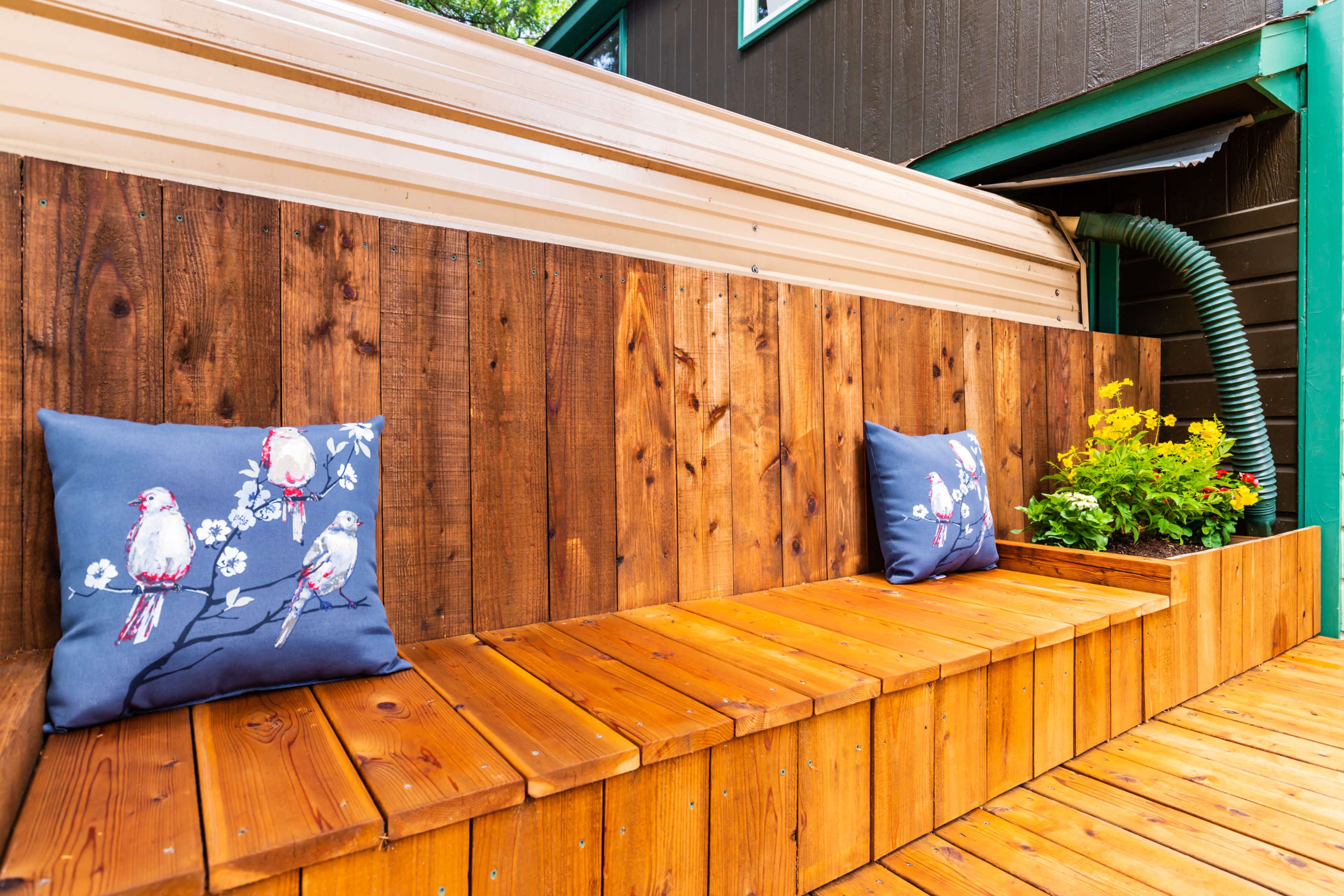 A wooden bench with two decorative pillows and a planter filled with flowers against a wooden wall.