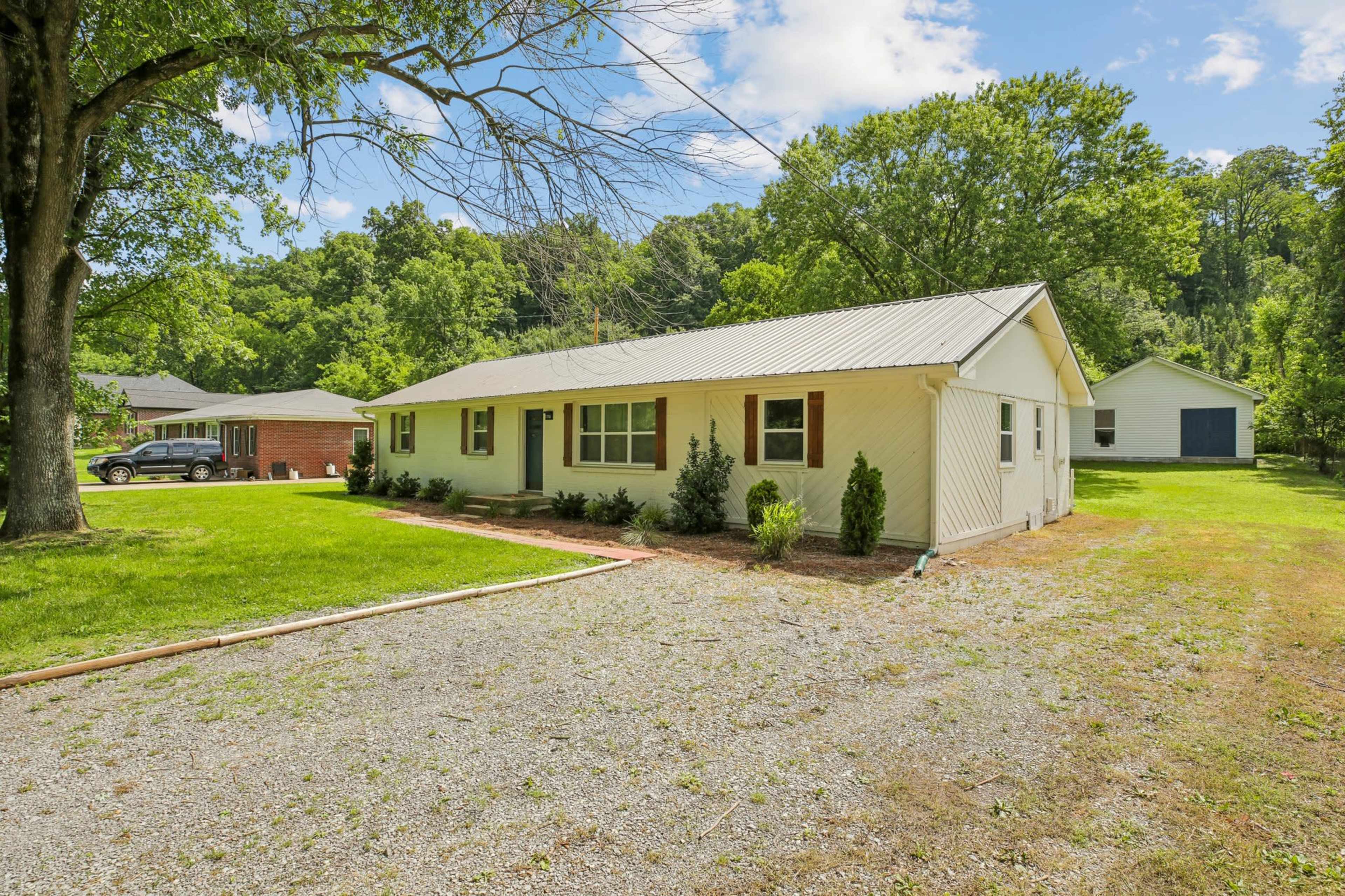 A single-story house with a metal roof and well-kept landscaping is situated on a gravel driveway, surrounded by green grass and trees.